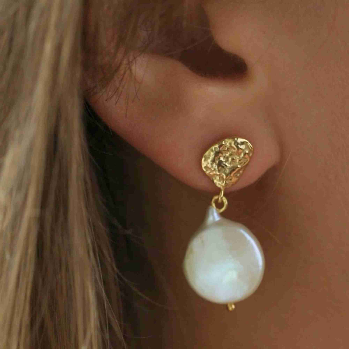 Macro close-up shot of a blonde model wearing the gold PEARLS GALORE Molten Silver & Keshi Pearl Drop Earrings, showcasing the hammered molten gold stud and organic flat white pearl drop.