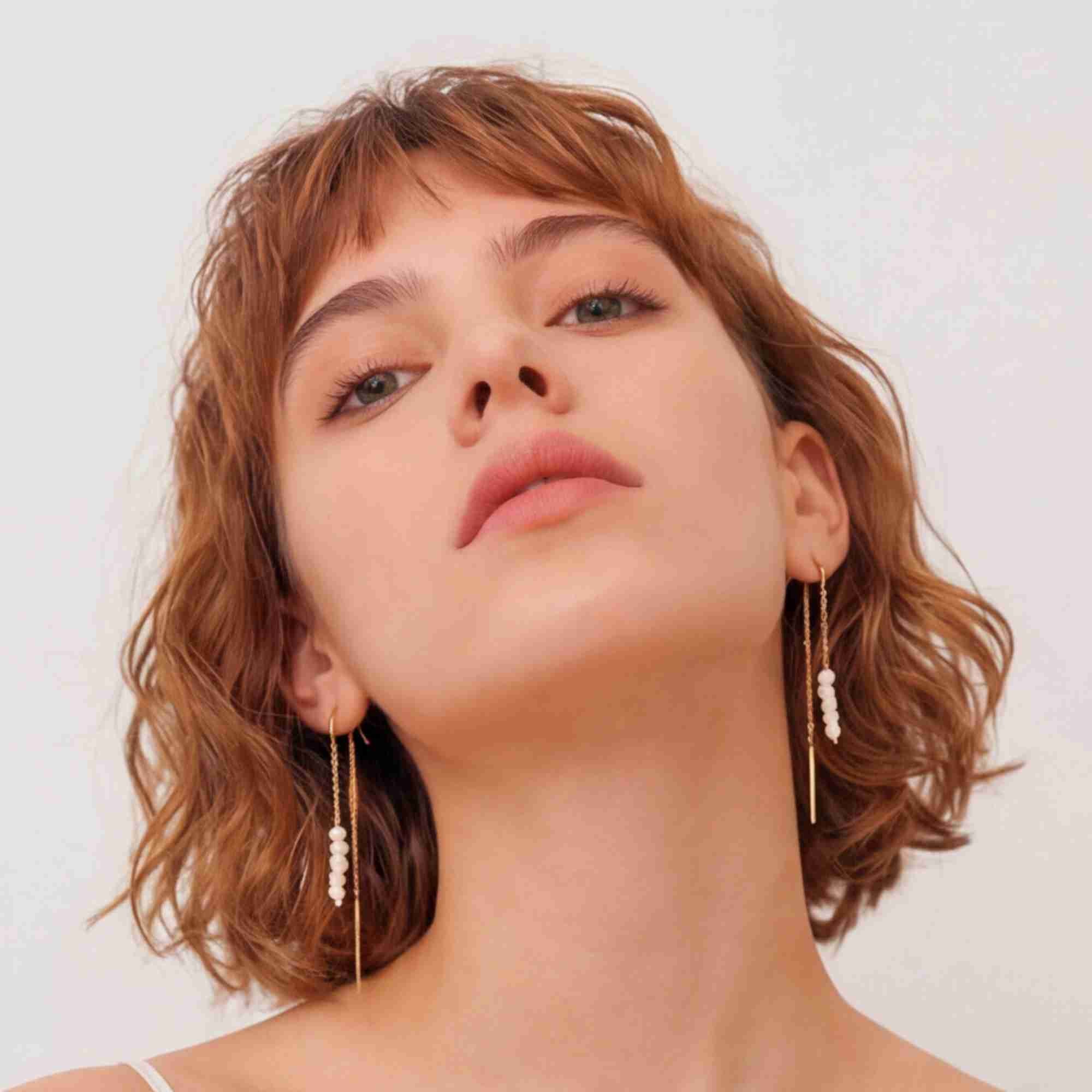Low-angle studio portrait of a model with a short brown wavy bob wearing the gold PEARLS GALORE Stacked Seed Pearl Chain Threader Earrings.