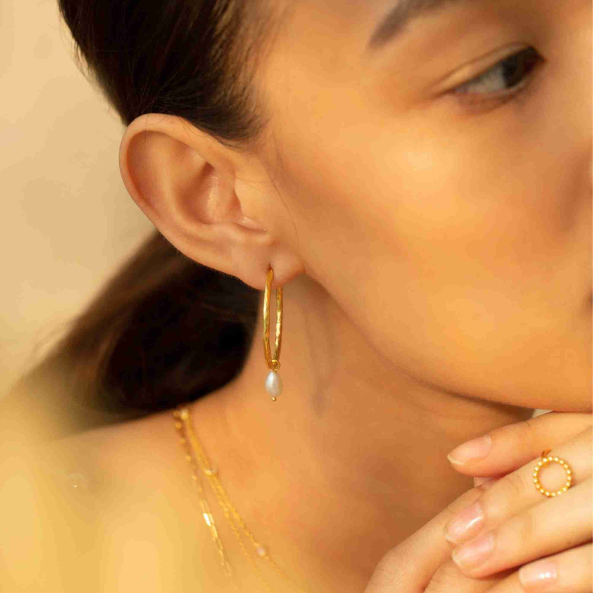 Three-quarter profile shot of a model wearing the gold pearl hoop earring paired with layered gold chain necklaces in warm sunlight.