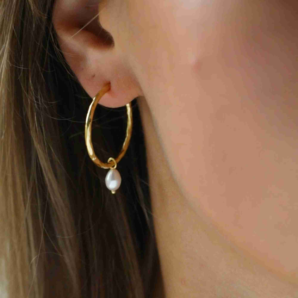 Extreme macro shot highlighting the textured gold hoop and dangling freshwater teardrop pearl charm worn on a model's earlobe.
