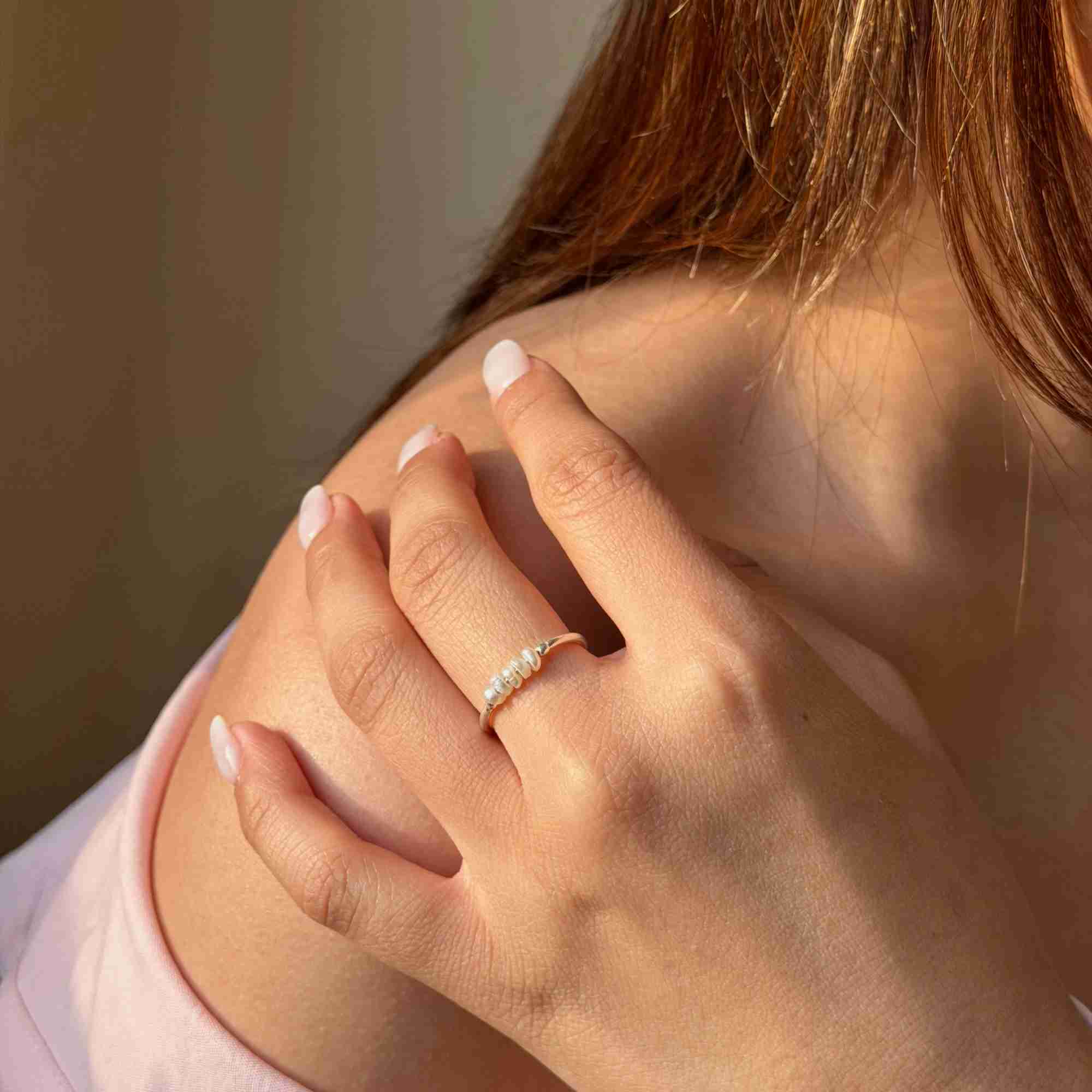 Warmly lit close-up of a model placing her hand on her shoulder, displaying the delicate silver band with a horizontal bar of small seed pearls.