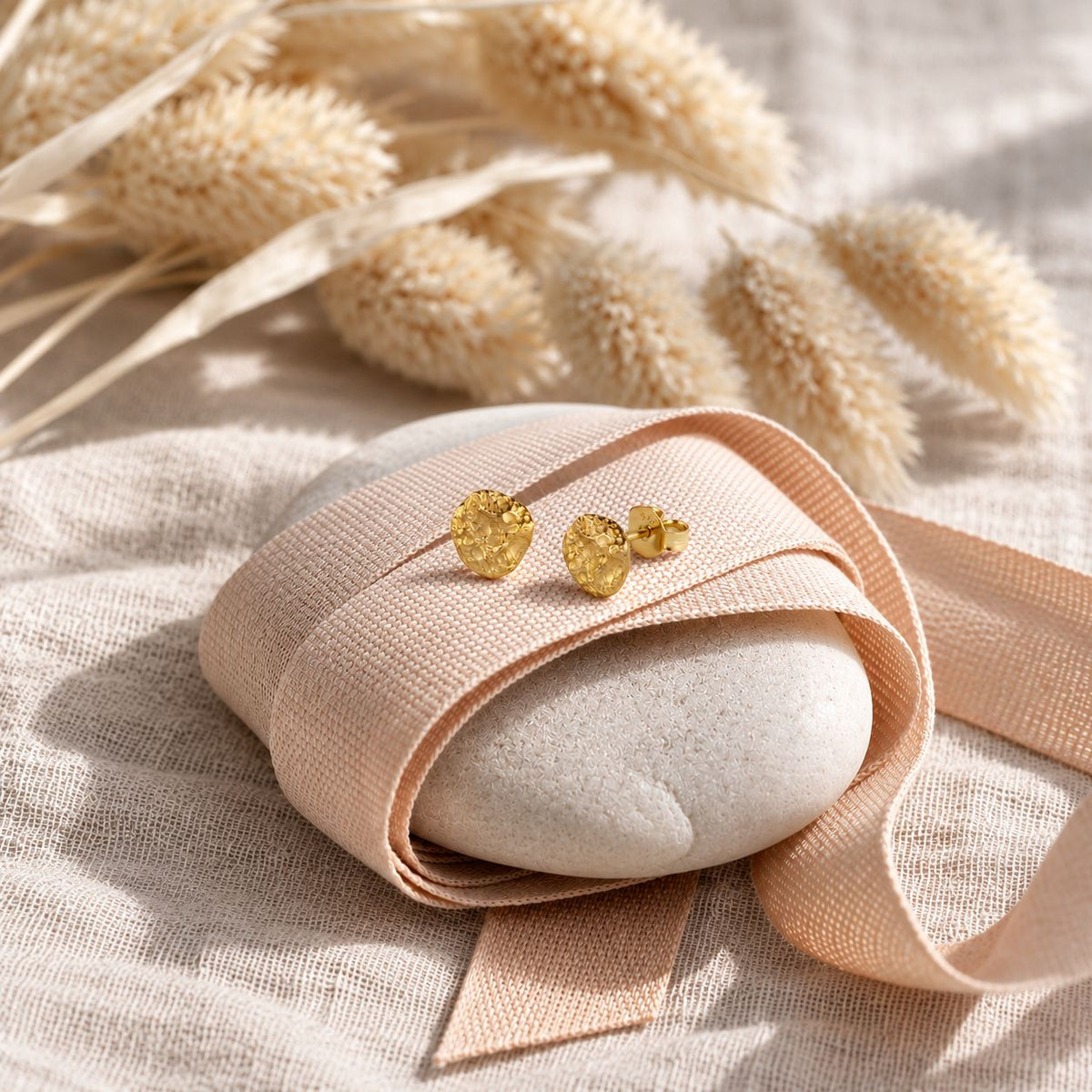 A pair of textured gold stud earrings displayed on a white stone wrapped in a peach ribbon, set on a beige linen fabric with dried bunny tail grass in the background.