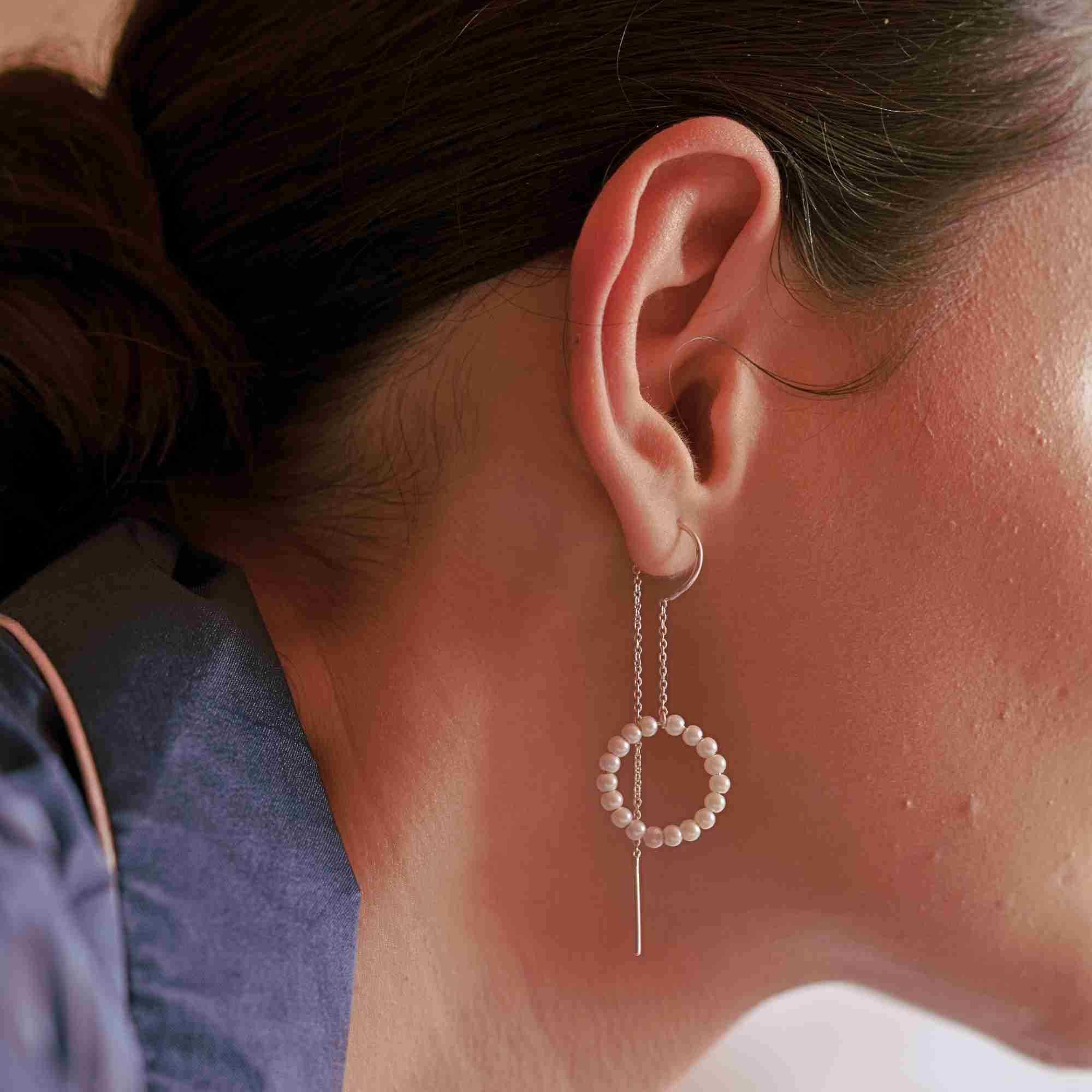 Close-up profile view of a model with dark hair in a bun wearing the silver Pearls Galore Seed Pearl Circle Chain Threader Earring, showcasing the delicate pearl circle pendant suspended from fine silver chains and a bar dangle.