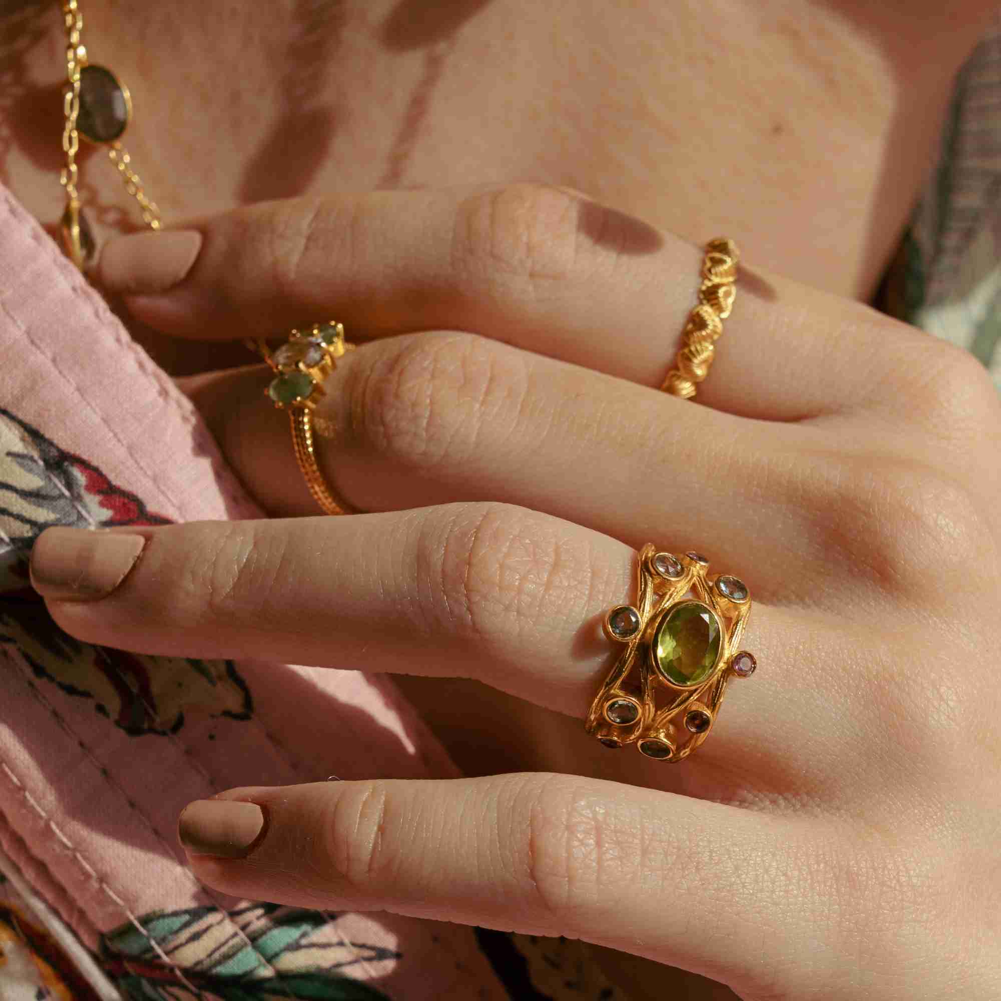 Warmly lit macro close-up of a model's hand with nude manicured nails, showing the gold Queen of Charms Fluted Hearts Eternity Band stacked alongside a statement gemstone ring and fine gold bands.