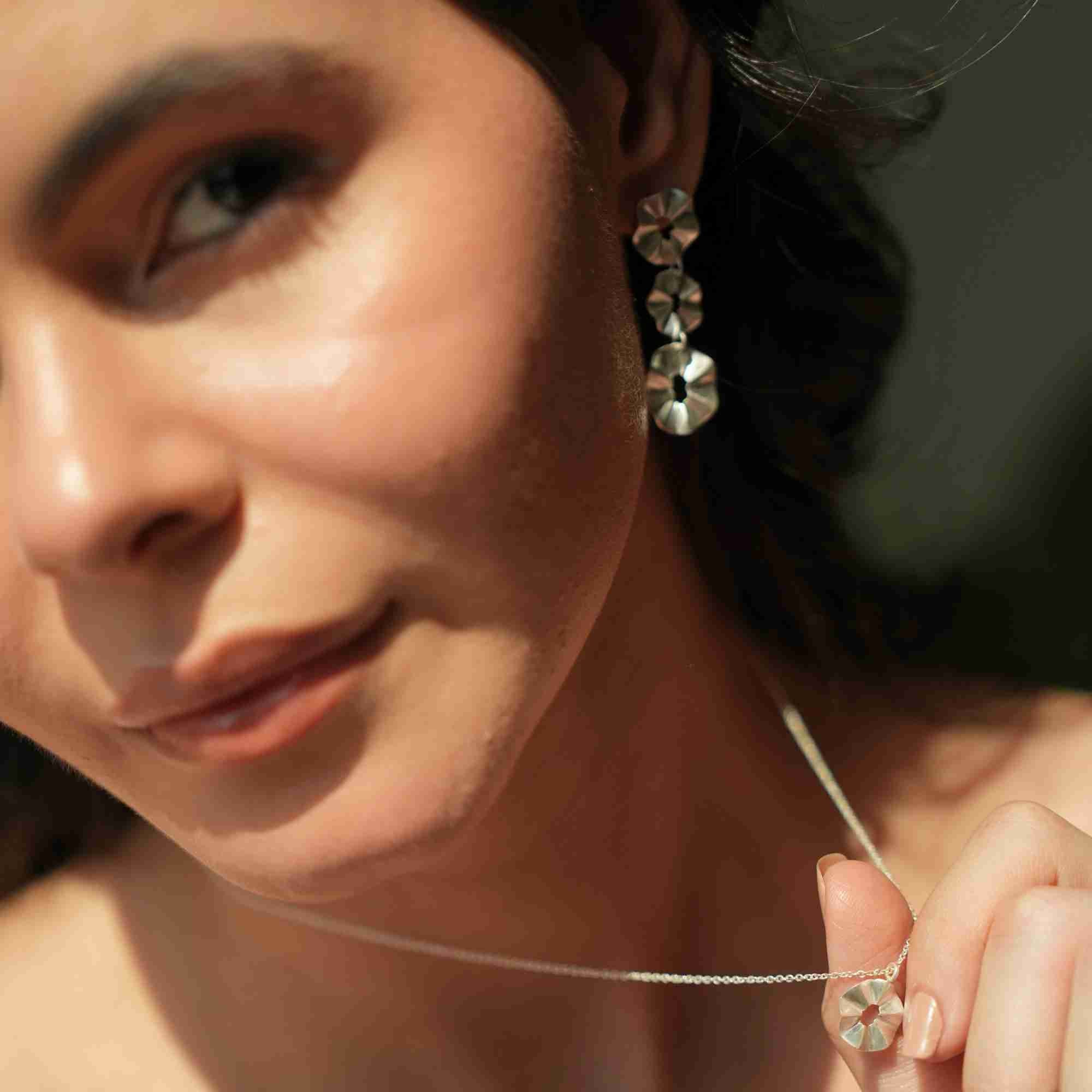 Close-up portrait of a model wearing the silver Ribbons & Lace Cascading Trio Statement Earrings, holding a matching silver flower pendant necklace in natural sunlight.