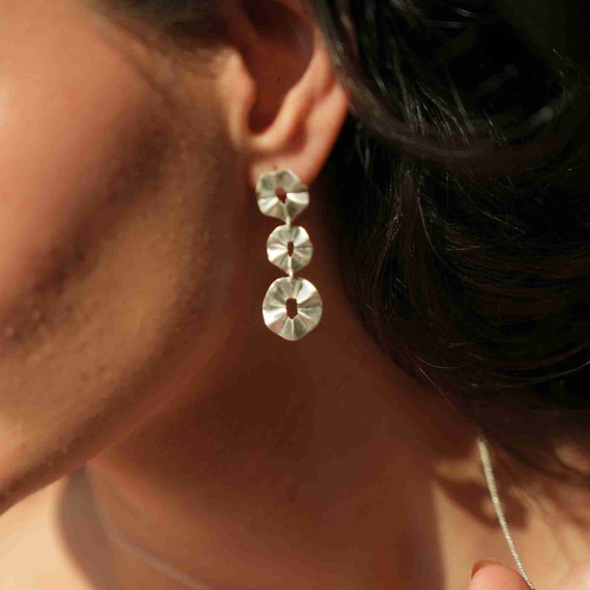 Warm-lit close-up profile view of a model wearing the sterling silver Ribbons & Lace Cascading Trio Statement Earrings, showcasing three cascading pleated disc shapes.