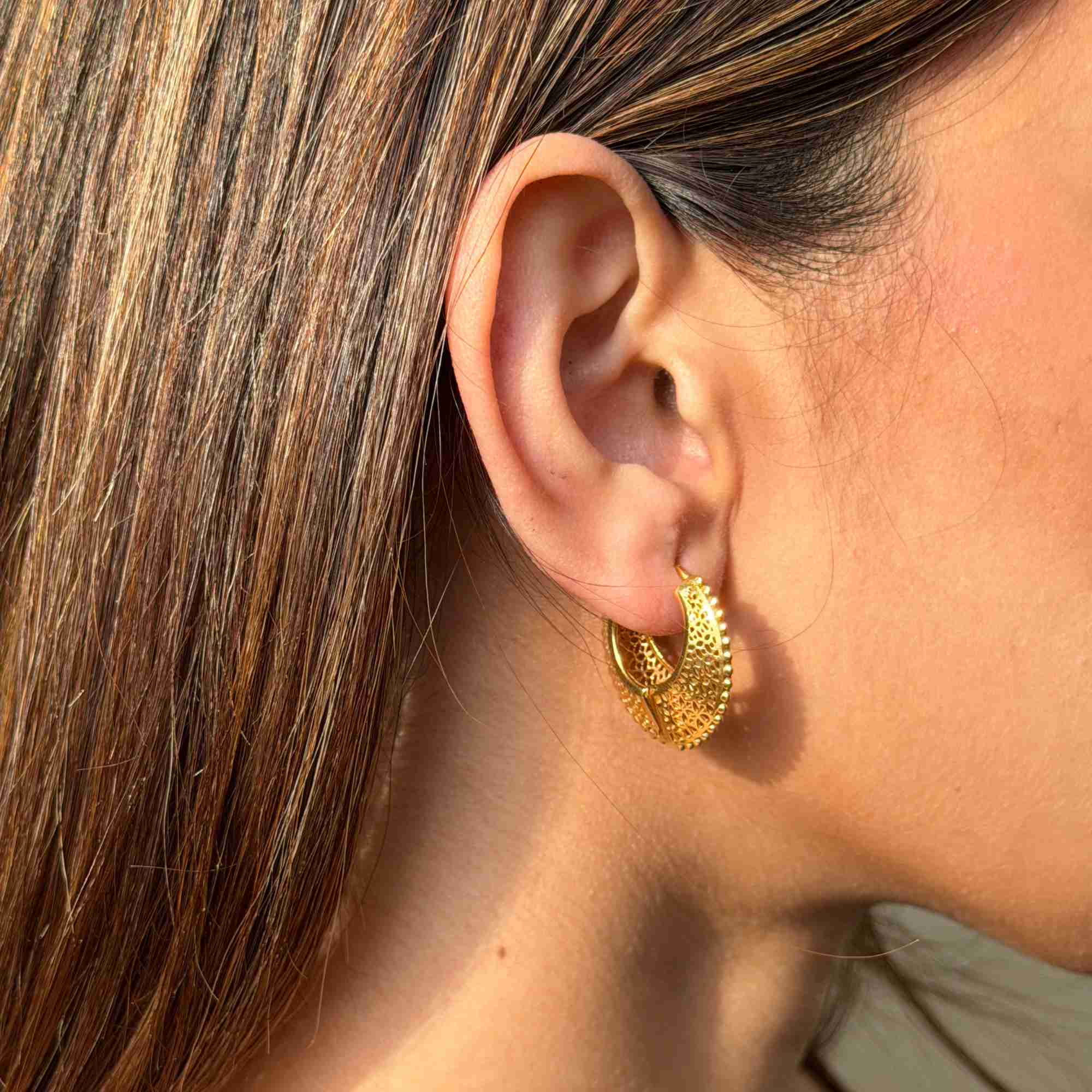 Macro side profile view highlighting the intricate openwork lace and filigree pattern on the gold Ribbons & Lace Sculptural Hoop Earrings worn on the model's ear.