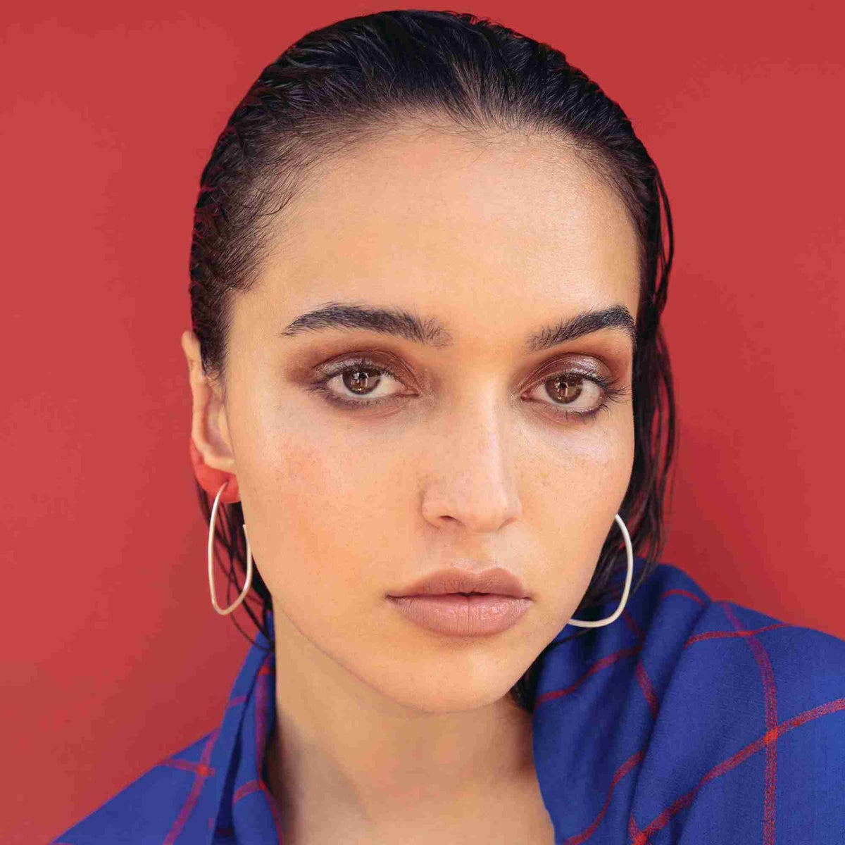 Editorial portrait against red, model with slicked-back hair wearing fine organic oval hoops in silver