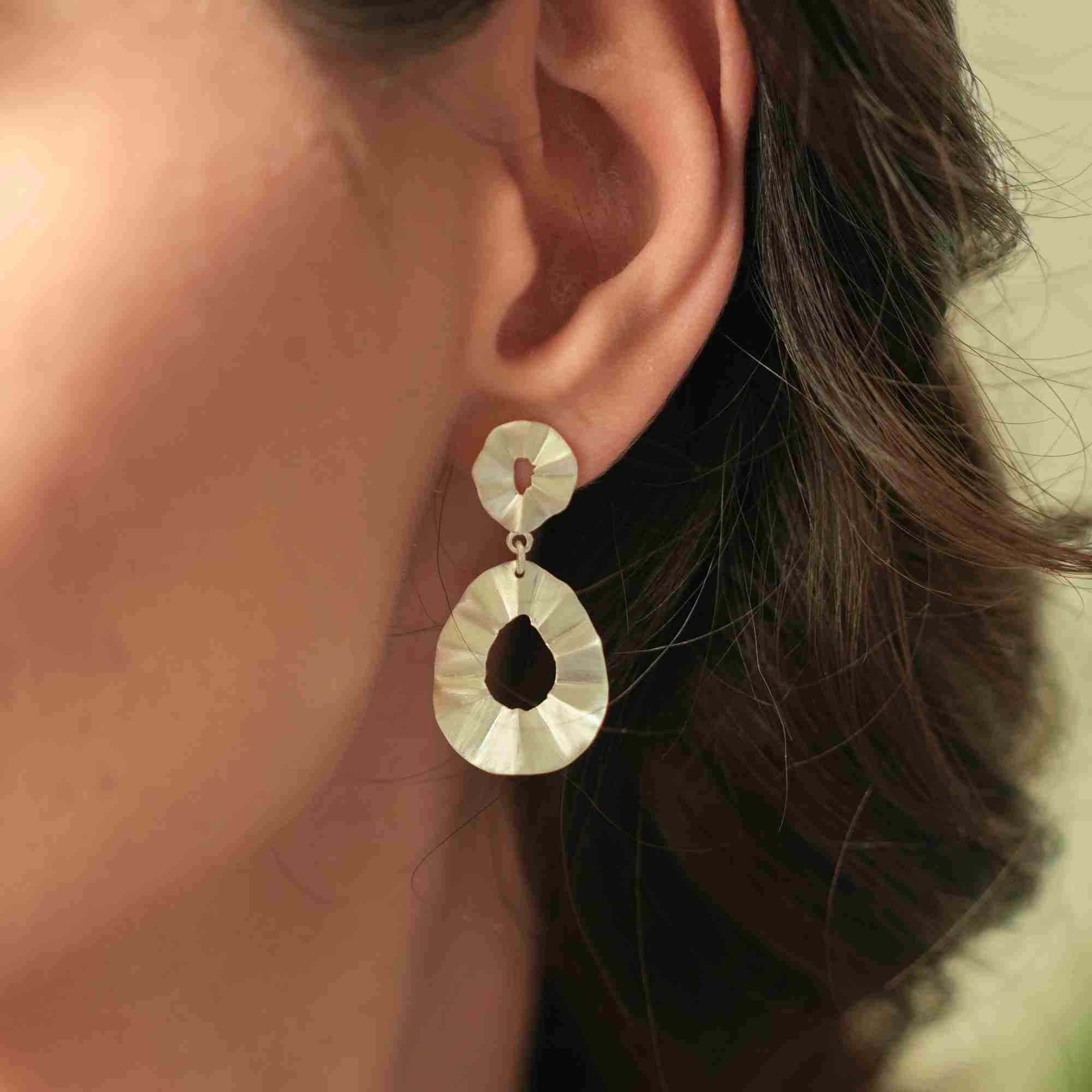 Warmly lit close-up shot of a model wearing the silver Ribbons & Lace Ruffle Fold Drop Statement Earrings, highlighting the double pleated disc drop design.
