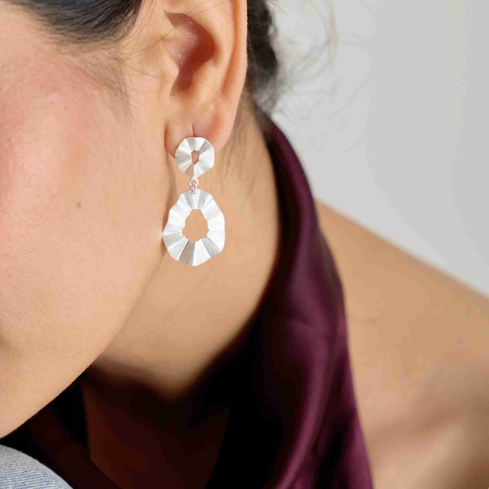 Side angle view named Ribbon & Lace showing a model wearing the Ribbons & Lace Ruffle Fold Drop Statement Earrings Silver, showcasing how the structured, sculptural matte silver folds catch the light against a deep burgundy top.