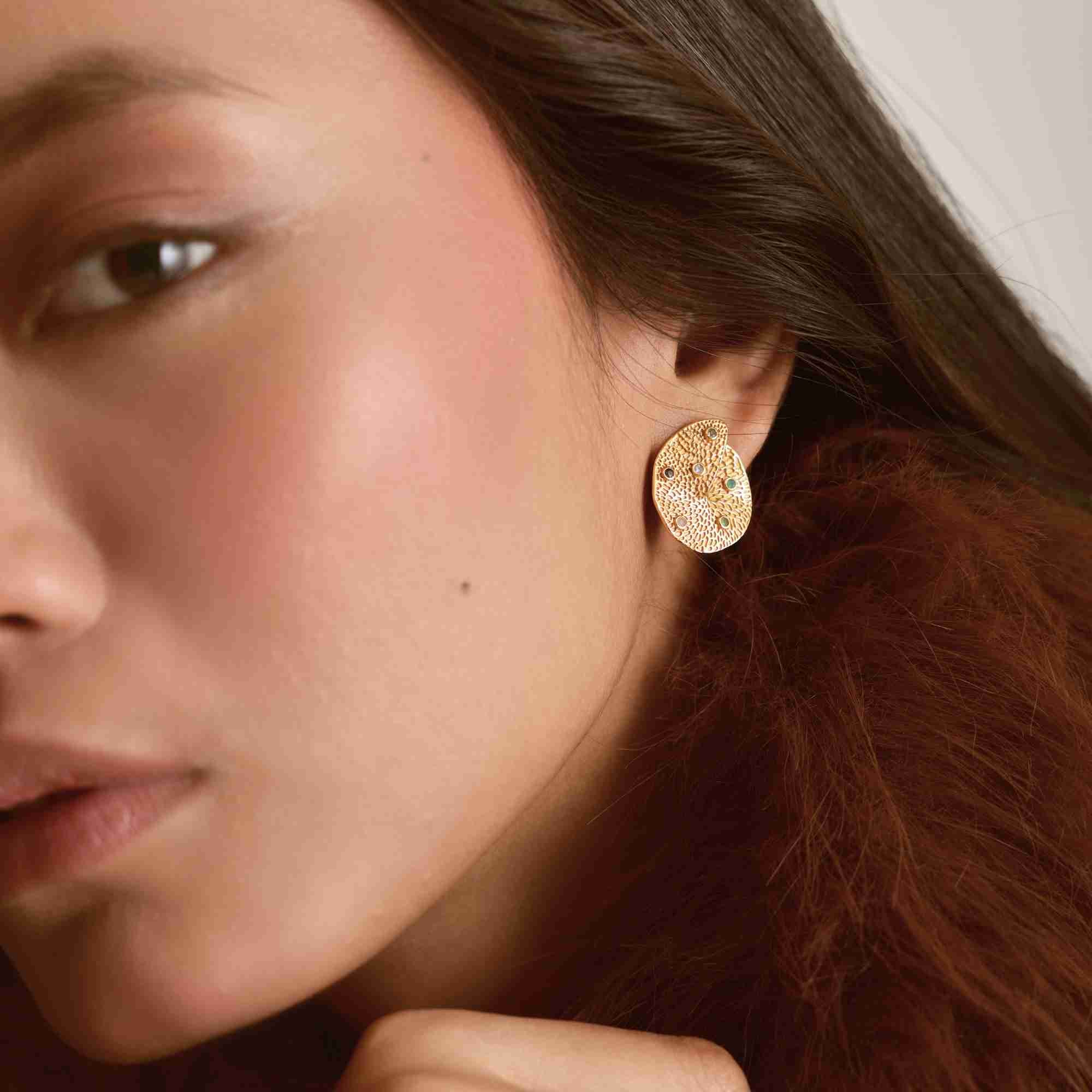 Angled portrait of a model showcasing the left earring, highlighting the hammer-textured gold surface studded with multi-colored crystals.
