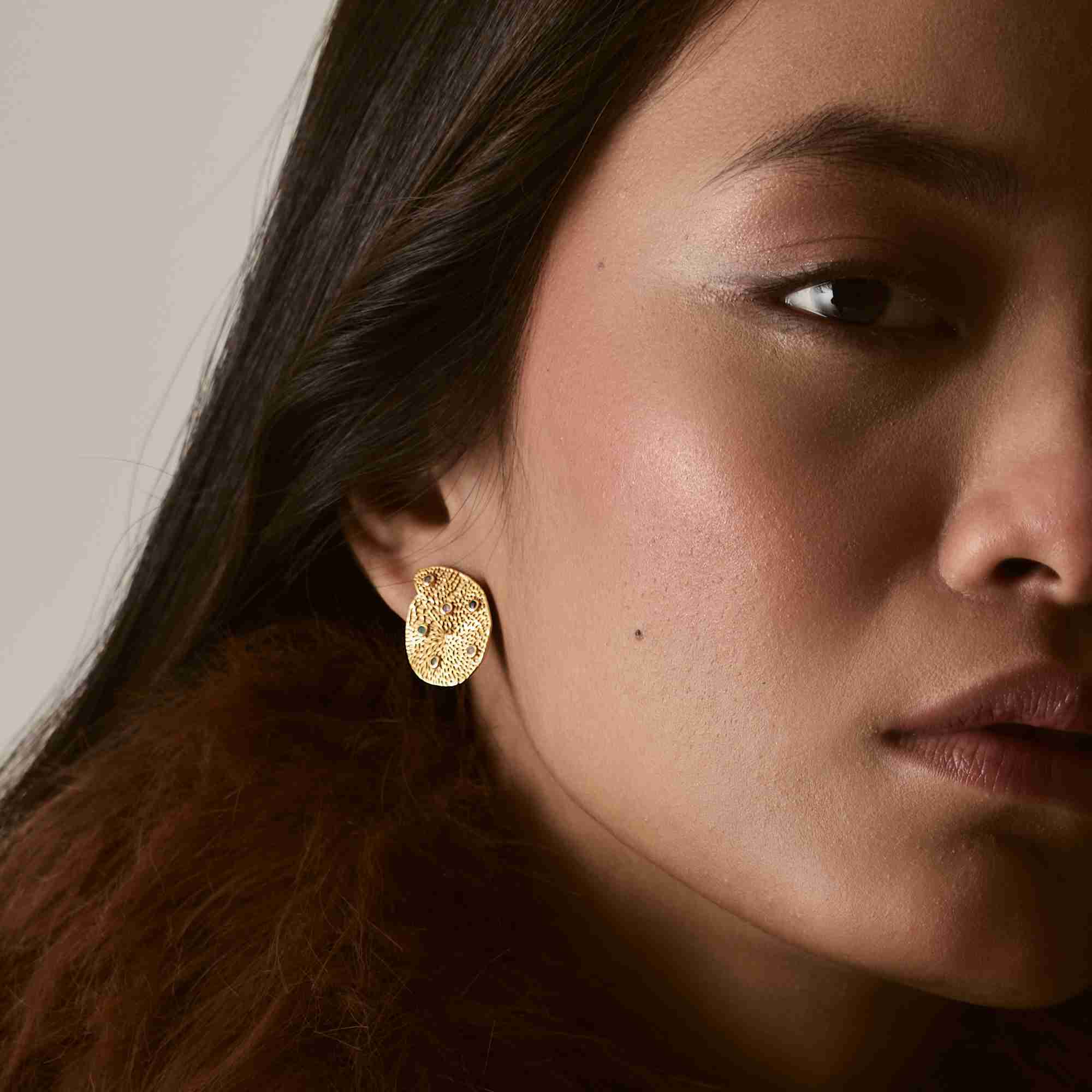 Close-up profile of a model wearing the gold Ribbons and Lace Galaxy Multi-Gemstone Stud Earrings, showing the textured disc inset with colorful tiny gemstones paired with a brown faux-fur collar.