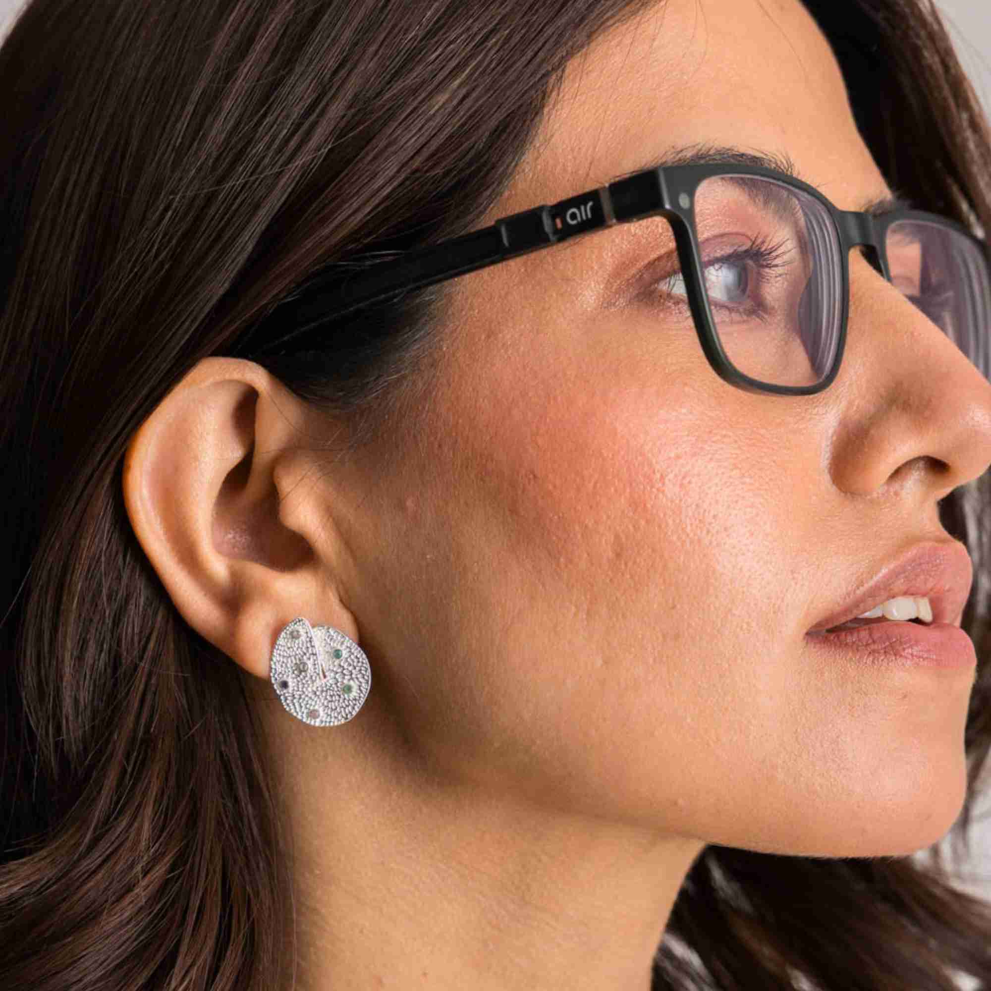 Side profile view of a model wearing black eyeglasses and the sterling silver Ribbons and Lace Galaxy Multi-Gemstone Stud Earrings, highlighting the textured silver disc inset with small colorful gemstones.