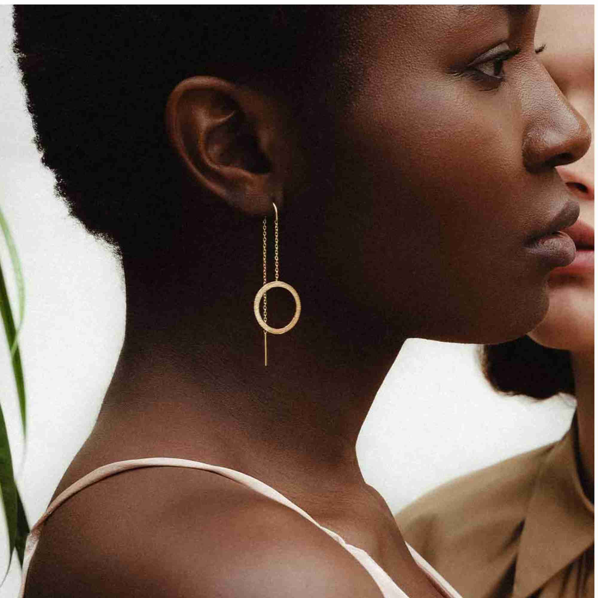 Studio product shot of Shimmering Disks Circle Chain Threader Earrings Gold lying flat on a textured white surface, showing the delicate box chain, sleek post bar, and dangling circular disc element with a brushed matte finish.