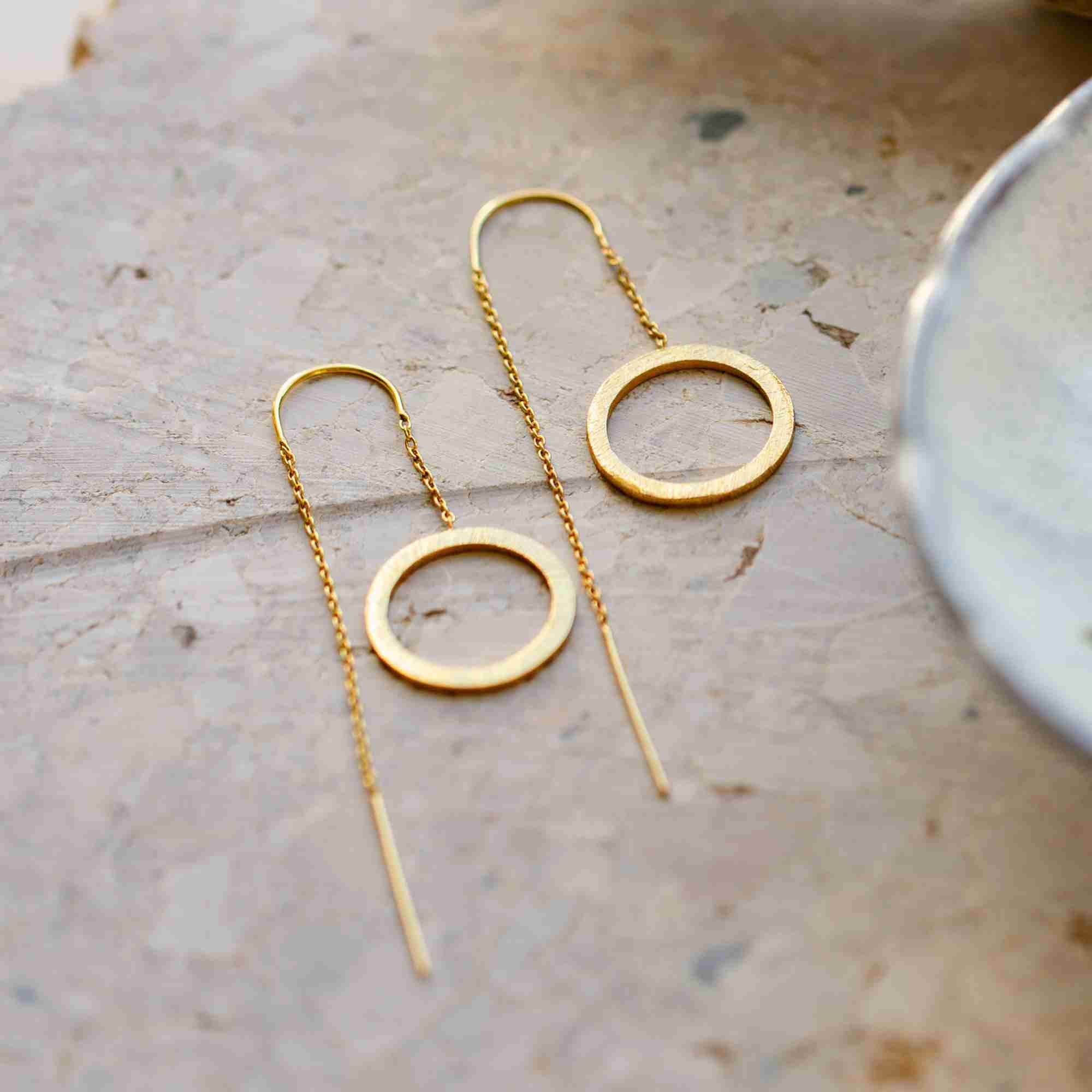 Flat lay photo of the pair of gold Shimmering Disks Circle Chain Threader Earrings resting on a light stone surface, displaying the full chain, bar end, and open circle pendant.