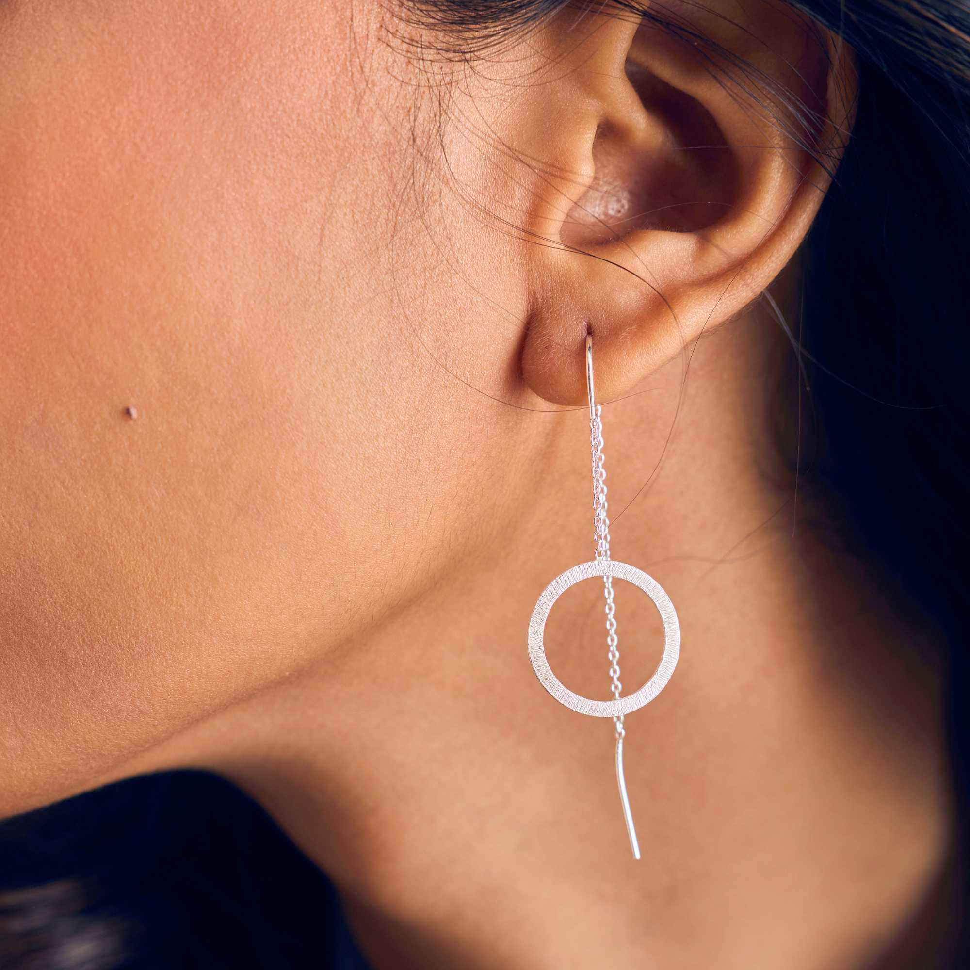 A model showcasing the Shimmering Disks Circle Chain Threader Earrings in silver, featuring a delicate fine drop chain that threads through the earlobe to suspend a textured, brushed metallic circular geometric charm.