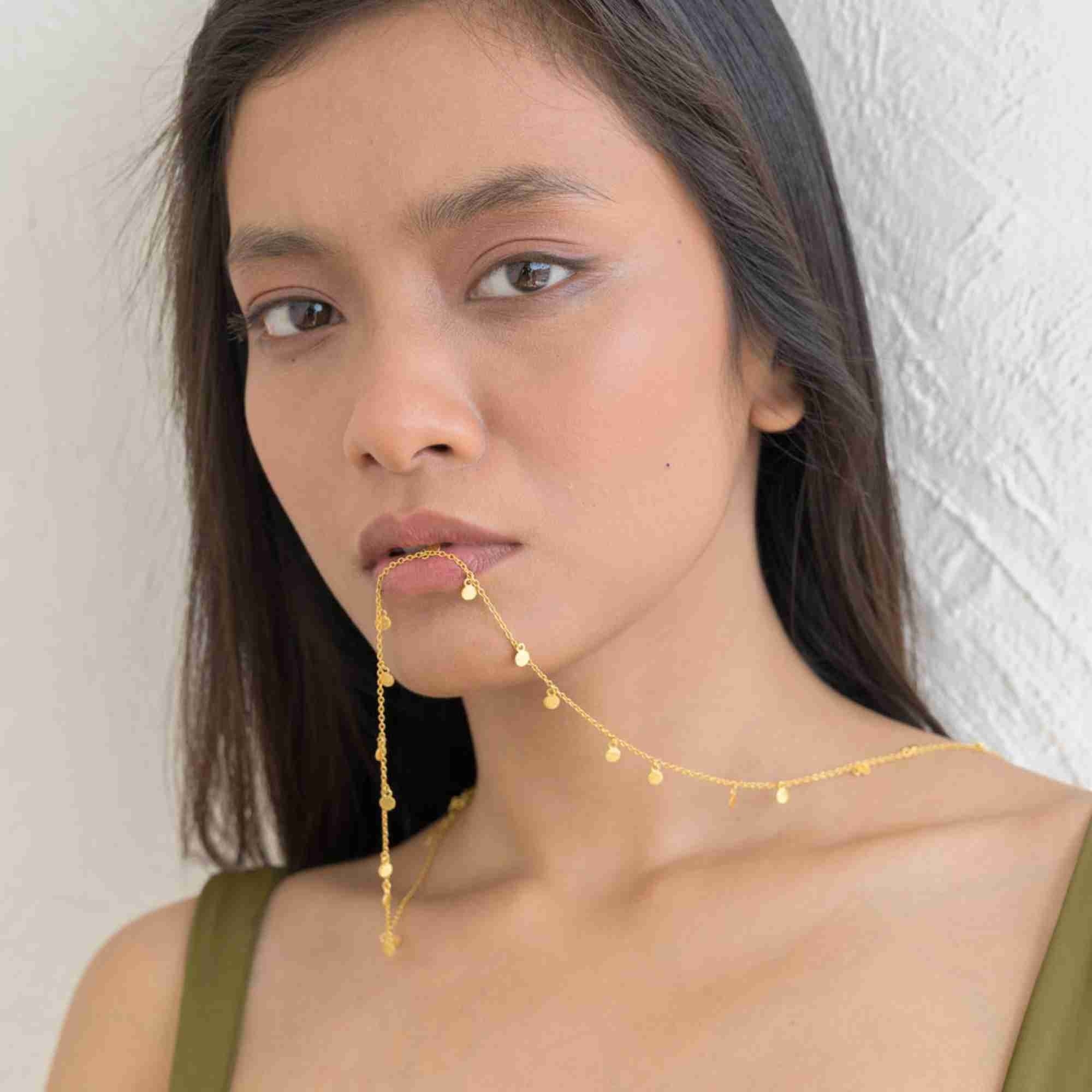A lifestyle close-up of a woman model holding the Shimmering Disks Constellation Disc Chain Necklace Gold in her mouth, displaying the delicate gold link chain and hanging mini disc details against a textured white background.