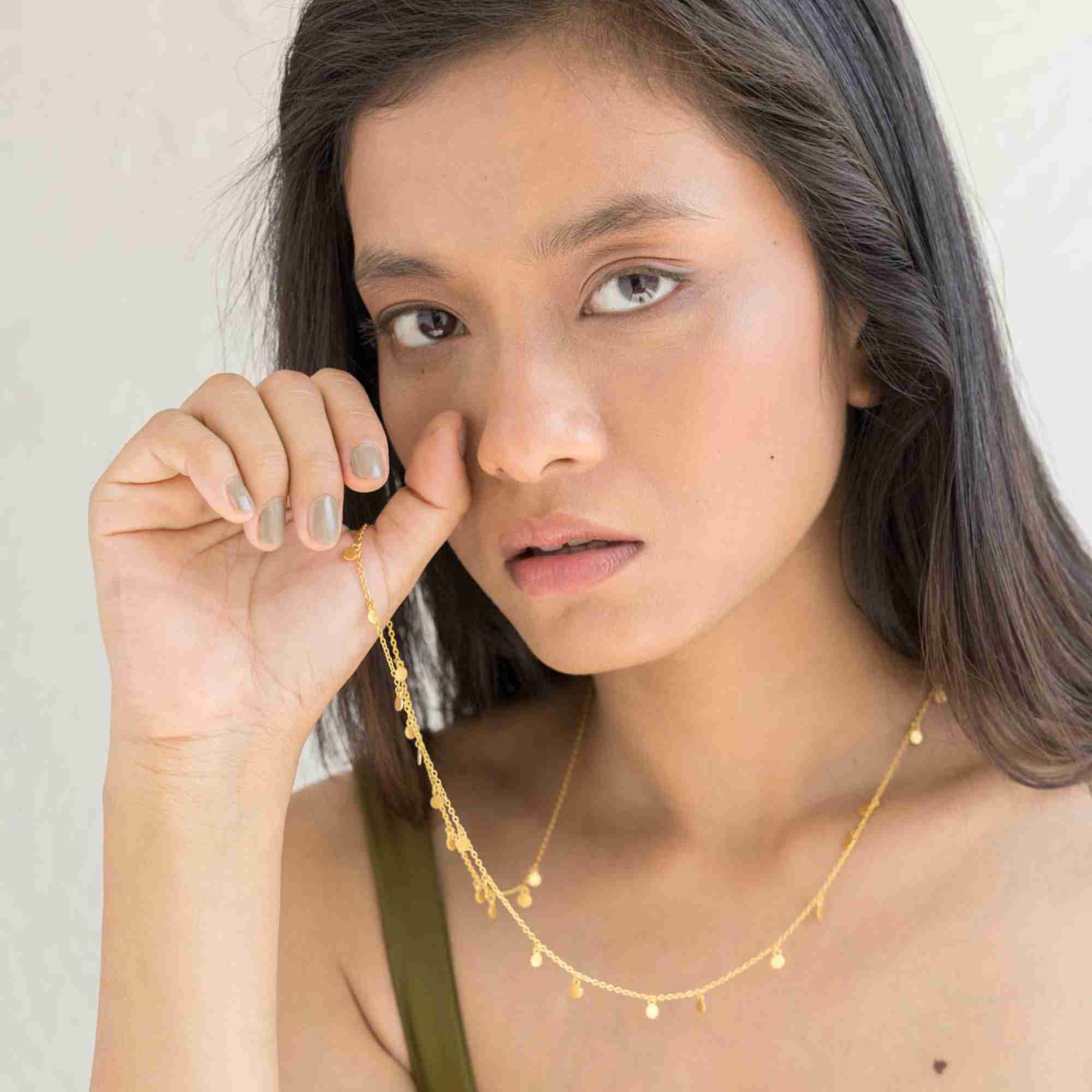 A lifestyle close-up of a woman model holding the Shimmering Disks Constellation Disc Chain Necklace Gold in her mouth, displaying the delicate gold link chain and hanging mini disc details against a textured white background.