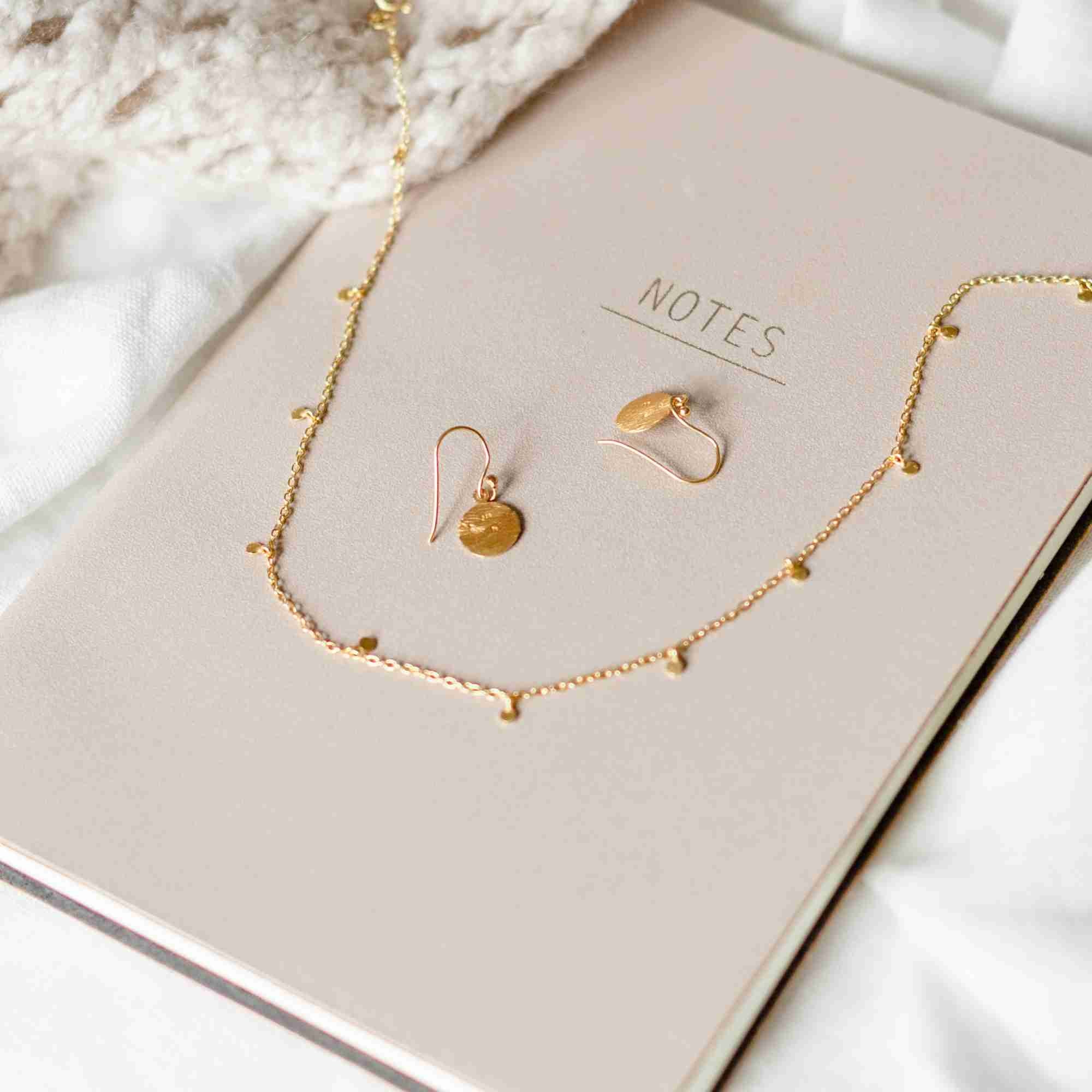 Studio product image named Shimmering Disks (57).jpg showing the Shimmering Disks Constellation Disc Chain Necklace Gold arranged around a pair of matching disc drop earrings on a beige "NOTES" journal; minimalist gold jewelry design by Protsaah Zurich.