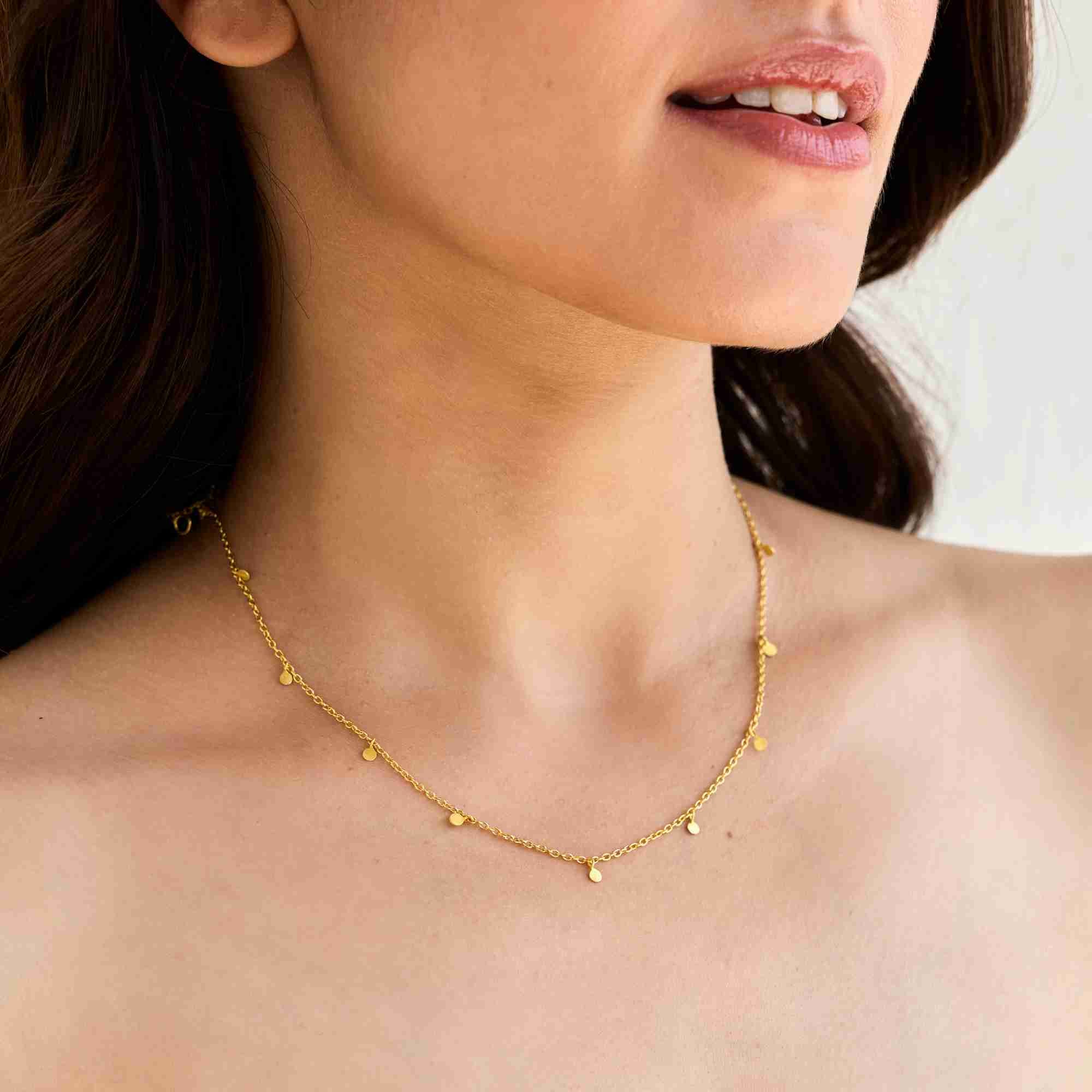 Gold Shimmering Disks Constellation Disc Chain Necklace laid out on a beige journal alongside matching gold drop earrings, displaying tiny coin charms spaced along the delicate chain.