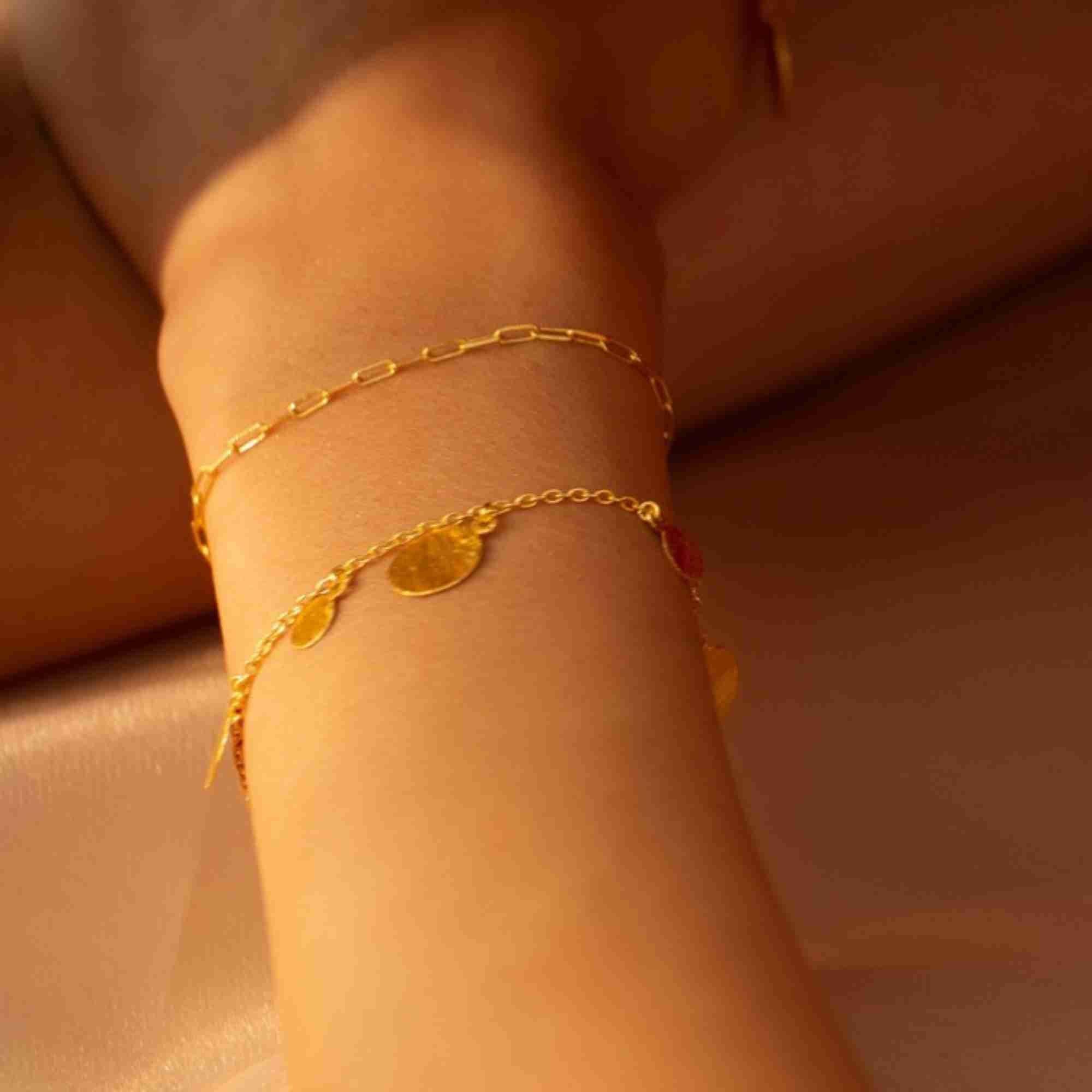 Close-up view of a model wearing two layered gold chain bracelets, featuring the Shimmering Disks Delicate Chain Bracelet with small dangling coin charms alongside a simple paperclip chain.