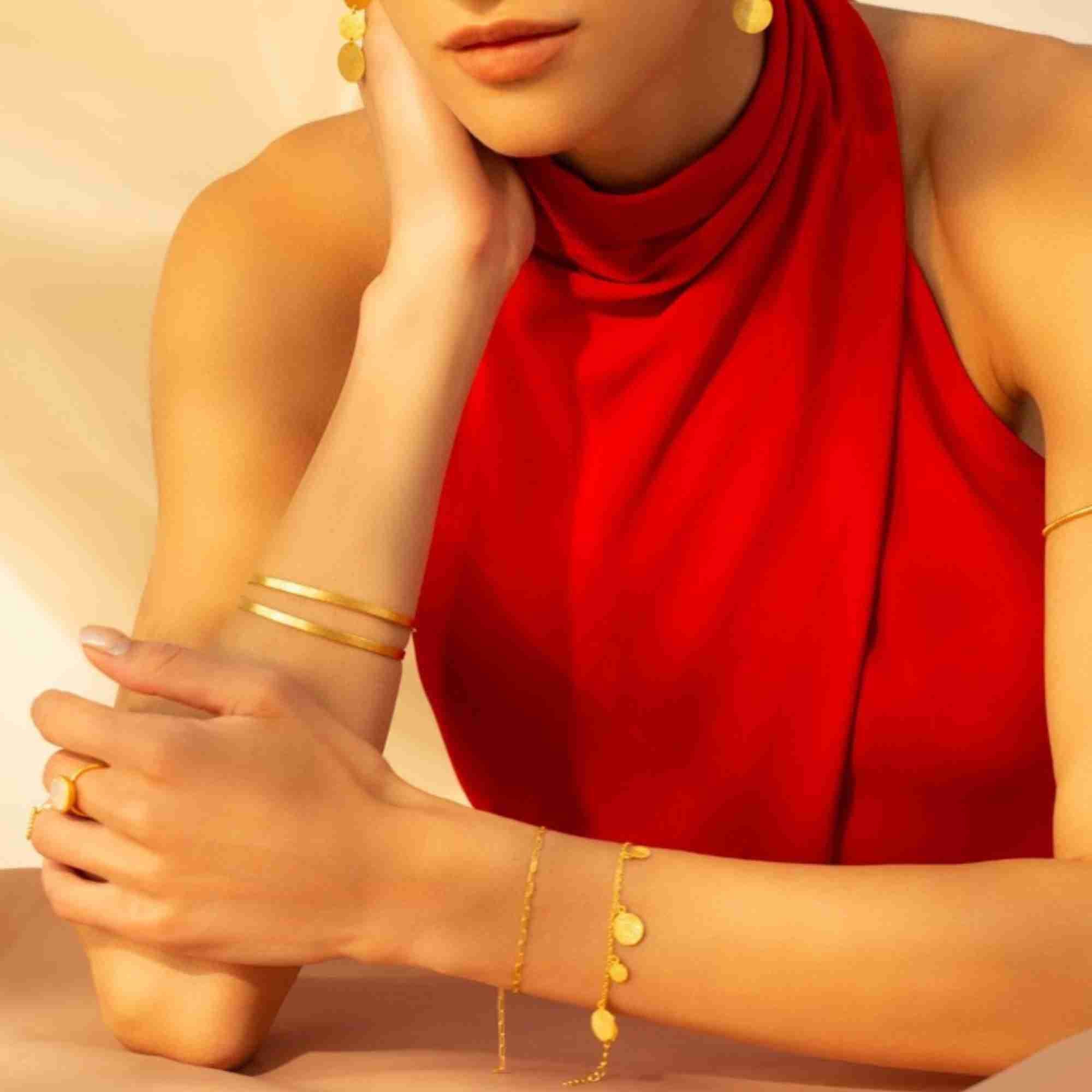 Model wearing a red halter top styled with the Shimmering Disks Delicate Chain Bracelet in gold, stacked with additional gold bangles and rings.