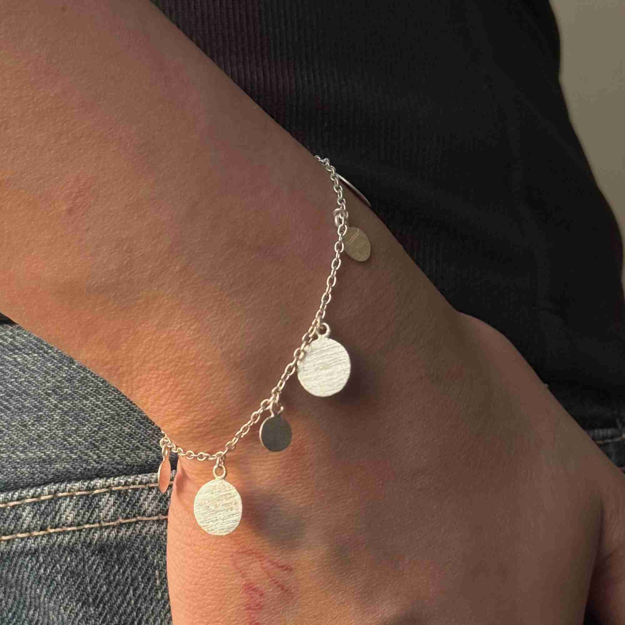 Close-up of the silver Shimmering Disks Delicate Chain Bracelet styled casually against dark clothing and denim jeans.