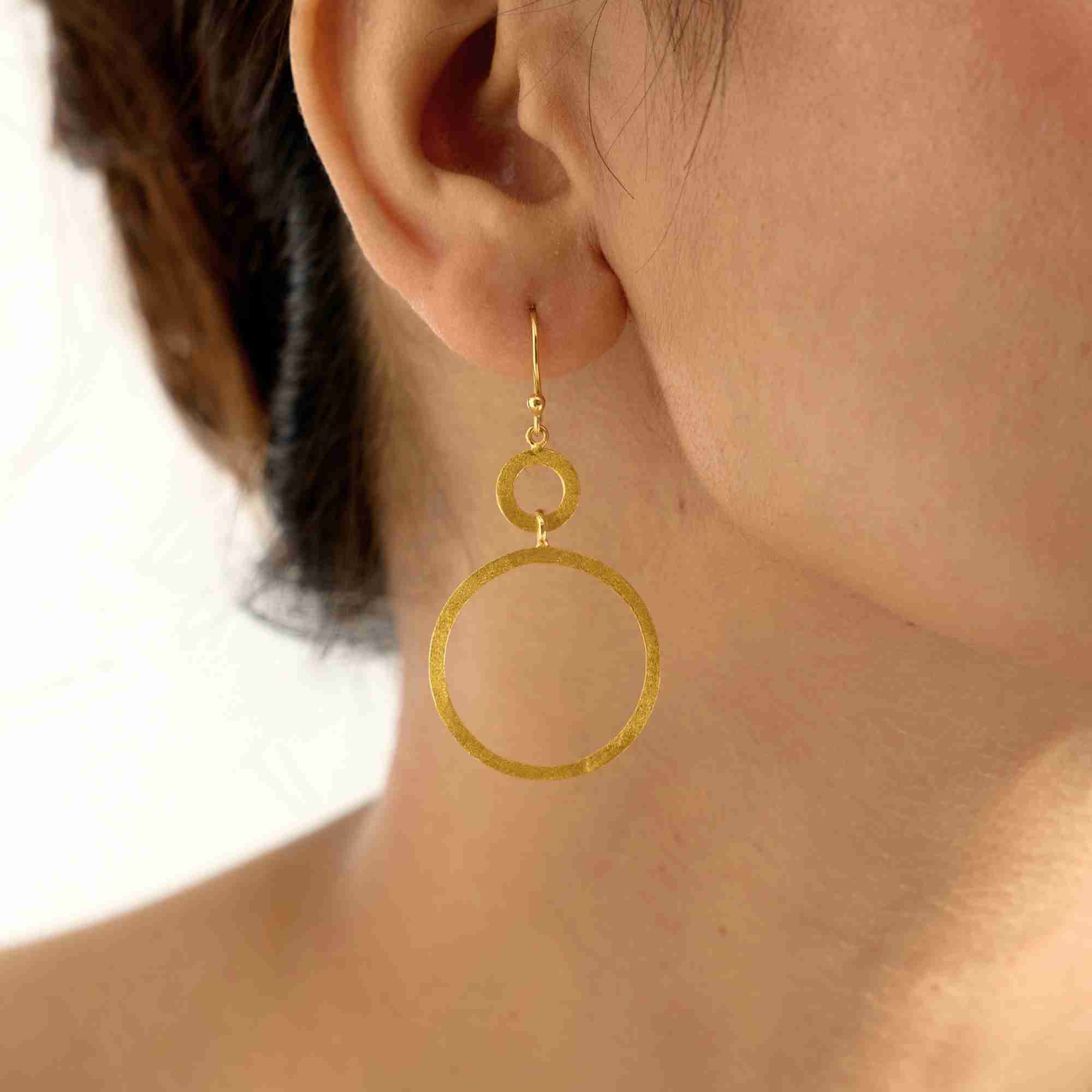 Macro detail shot of the gold Shimmering Disks Dual Circle Statement Earring, highlighting the hand-brushed finish on the small top ring and larger bottom ring.