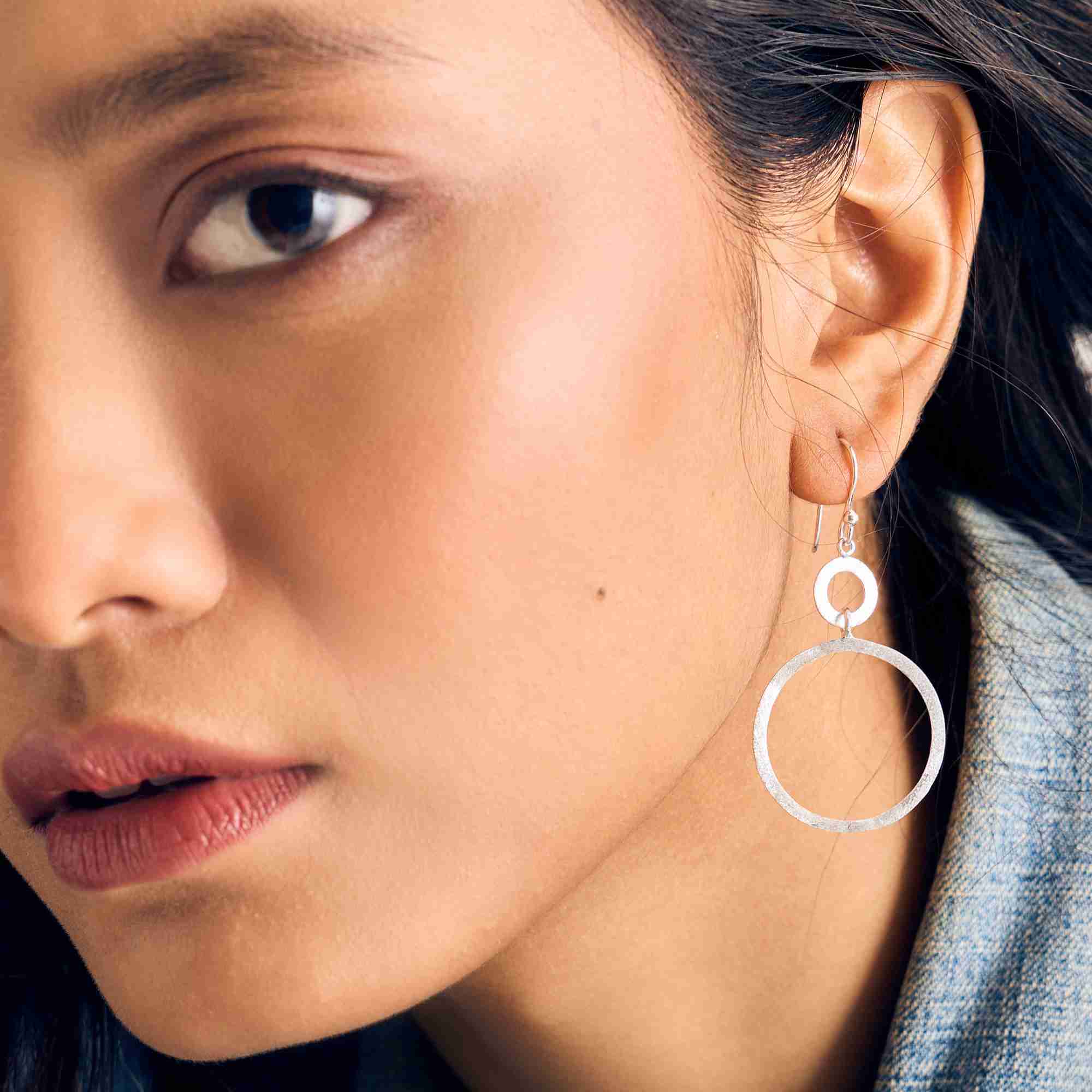Front-angled close-up portrait of a model wearing the silver Shimmering Disks Dual Circle Statement Earring, paired with a denim jacket.
