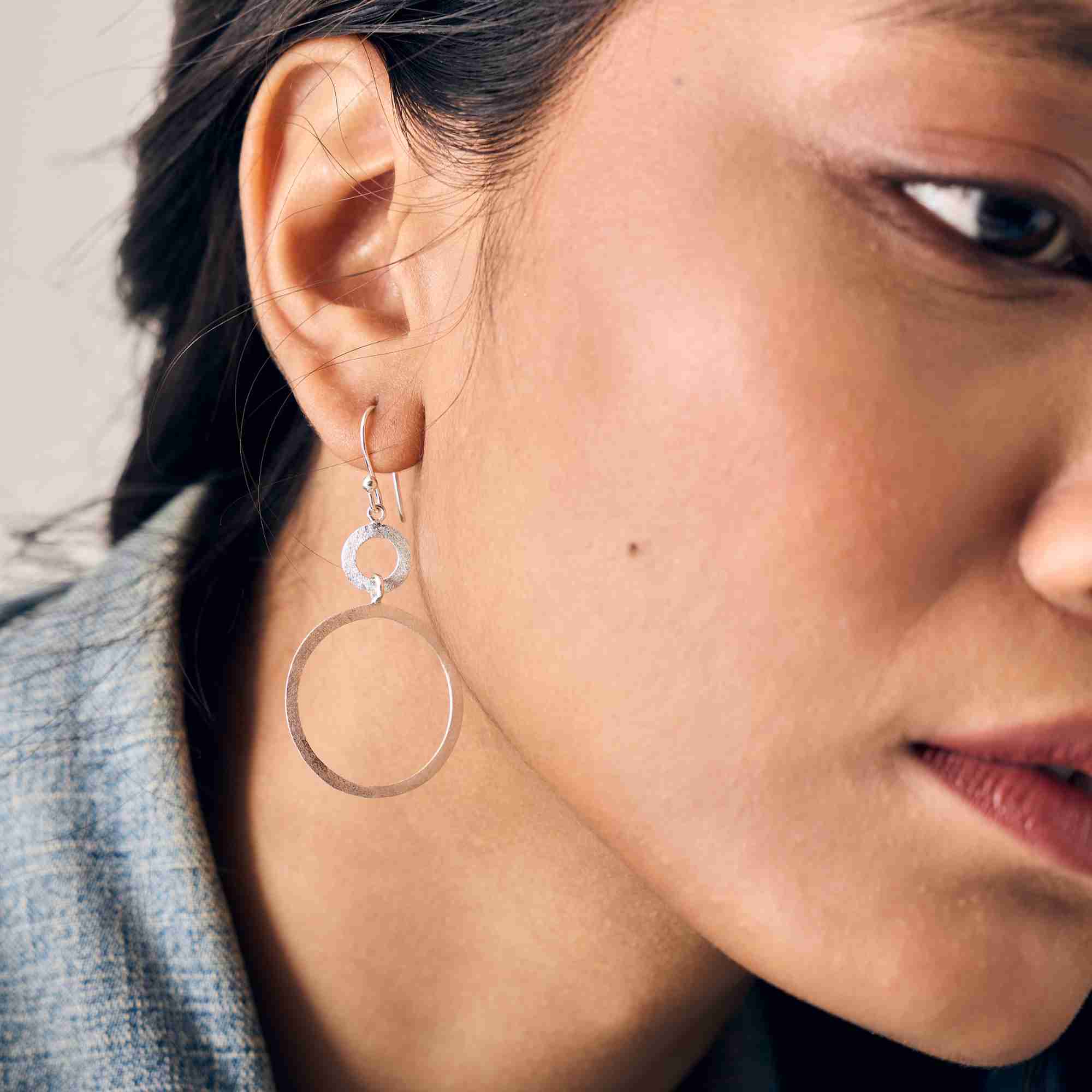 Macro shot of a model wearing the Shimmering Disks Dual Circle Statement Earring in silver, featuring a small textured ring linked to a larger open circle on a french wire hook.