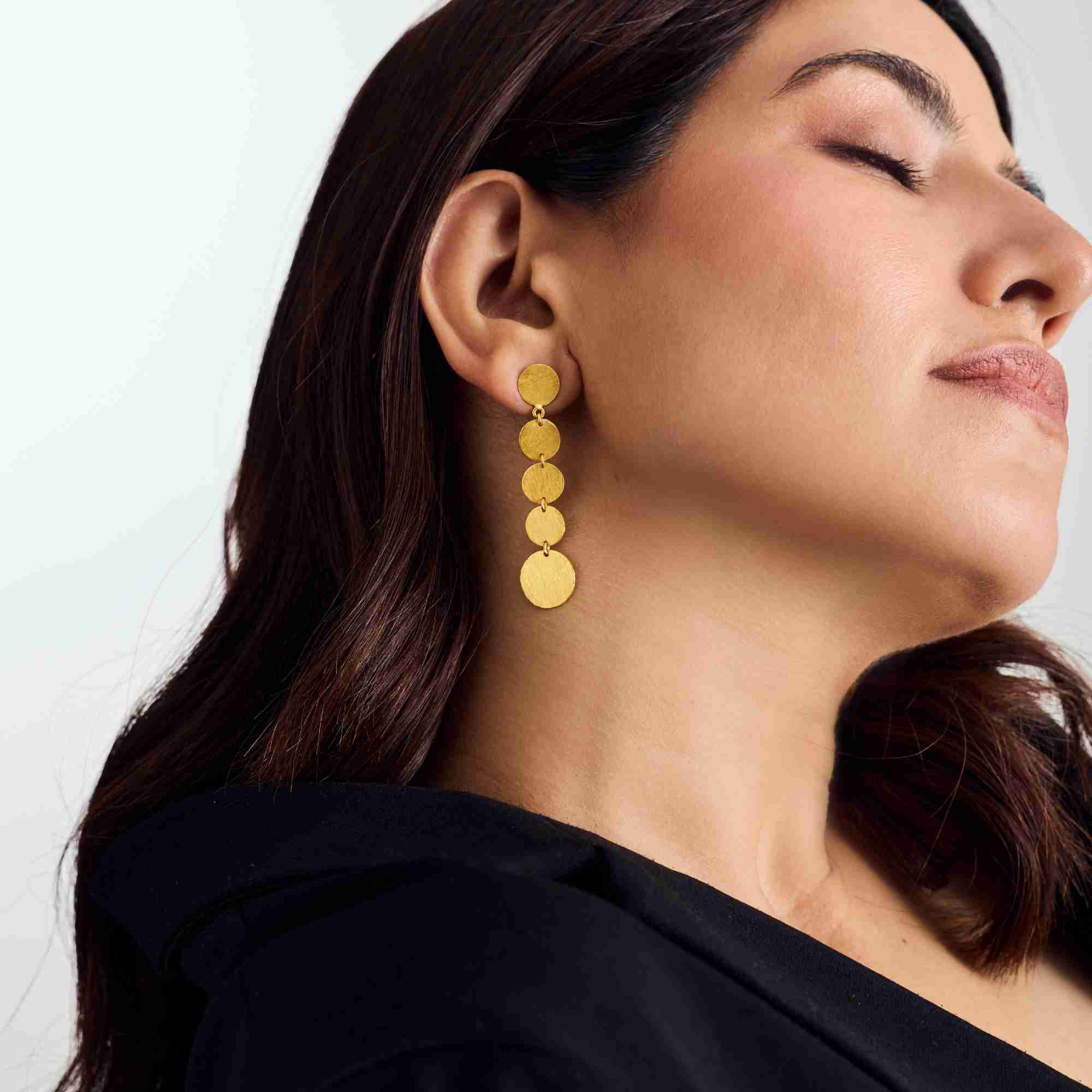 Studio portrait of a model looking upward while wearing the gold Shimmering Disks Graduating Disc Cascade Statement Earrings.