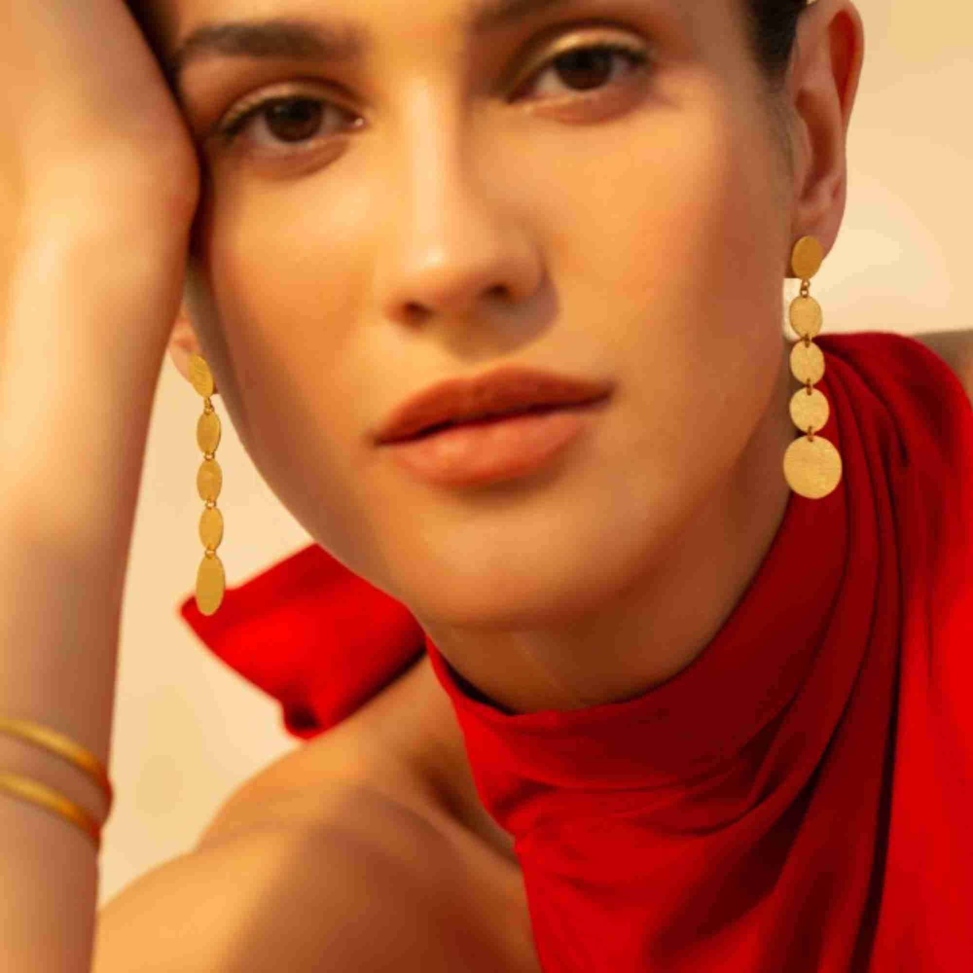 Warm-toned close-up portrait of a model wearing gold Shimmering Disks Graduating Disc Cascade Statement Earrings in a red halter top.