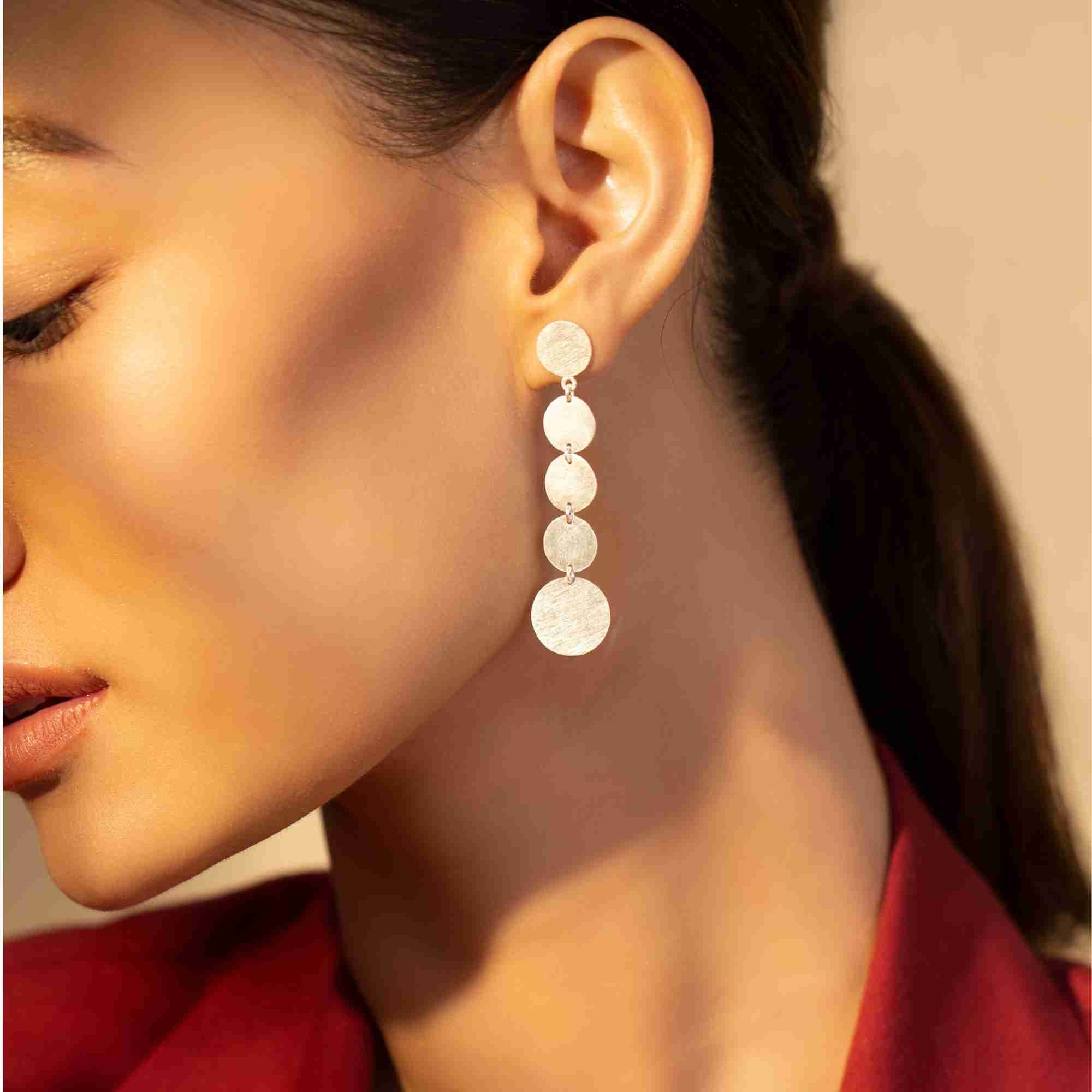 Side profile of a model wearing the silver Shimmering Disks Graduating Disc Cascade Statement Earring, showcasing five brushed-finish circular discs linked in a cascading drop design.