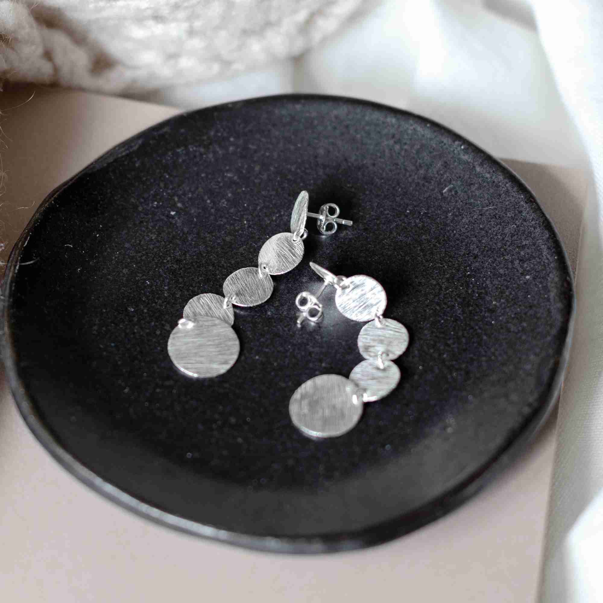 Pair of silver Shimmering Disks Graduating Disc Cascade Statement Earrings with post backs lying on a small, dark grey ceramic dish.