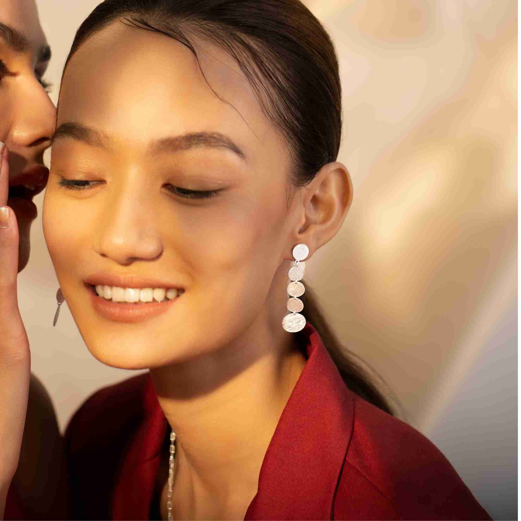 Smiling model wearing the silver Shimmering Disks Graduating Disc Cascade Statement Earring in warm light, paired with a red blazer.