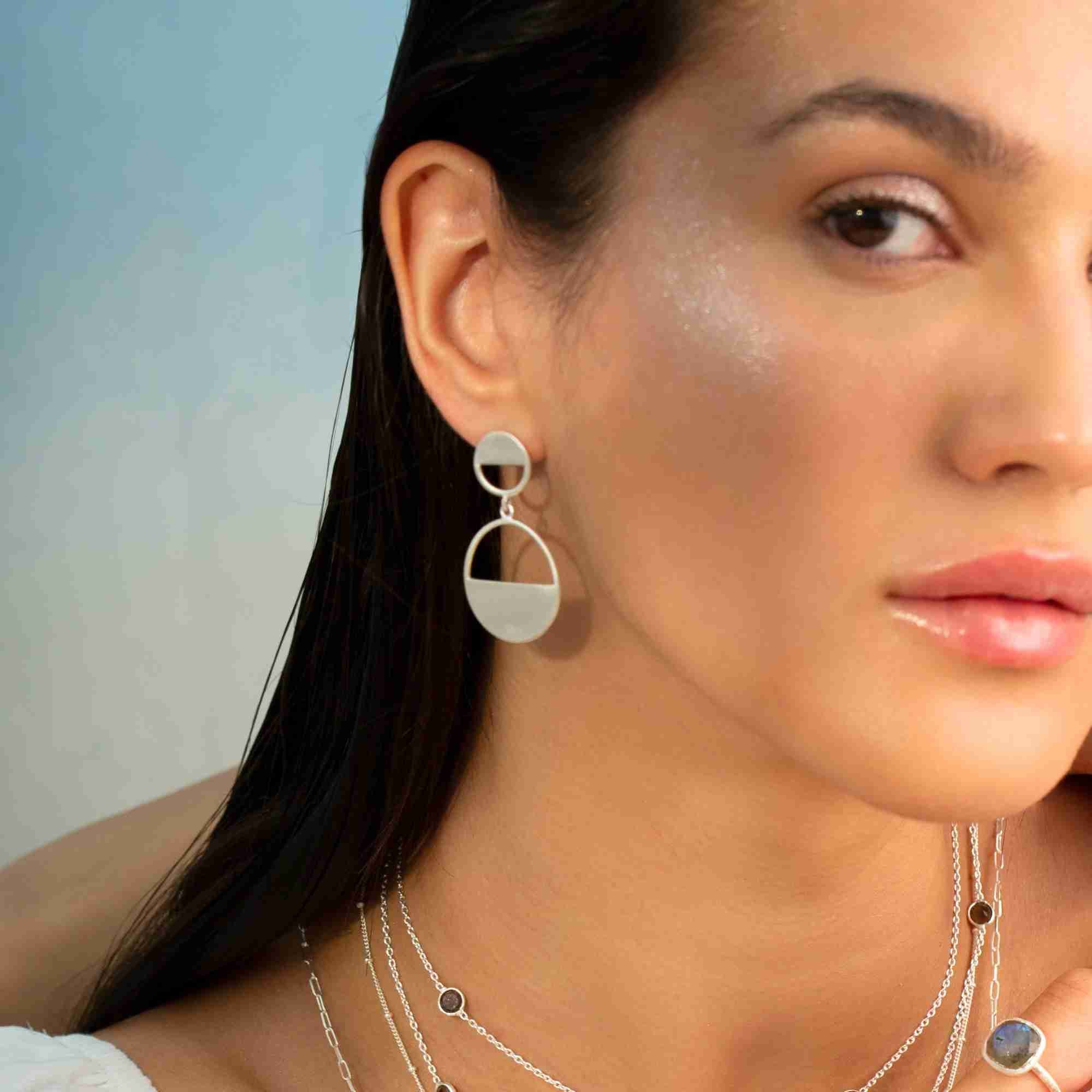 Close-up portrait of a model wearing the silver Shimmering Disks Horizon Duo Drop Statement Earring, paired with layered silver necklaces.