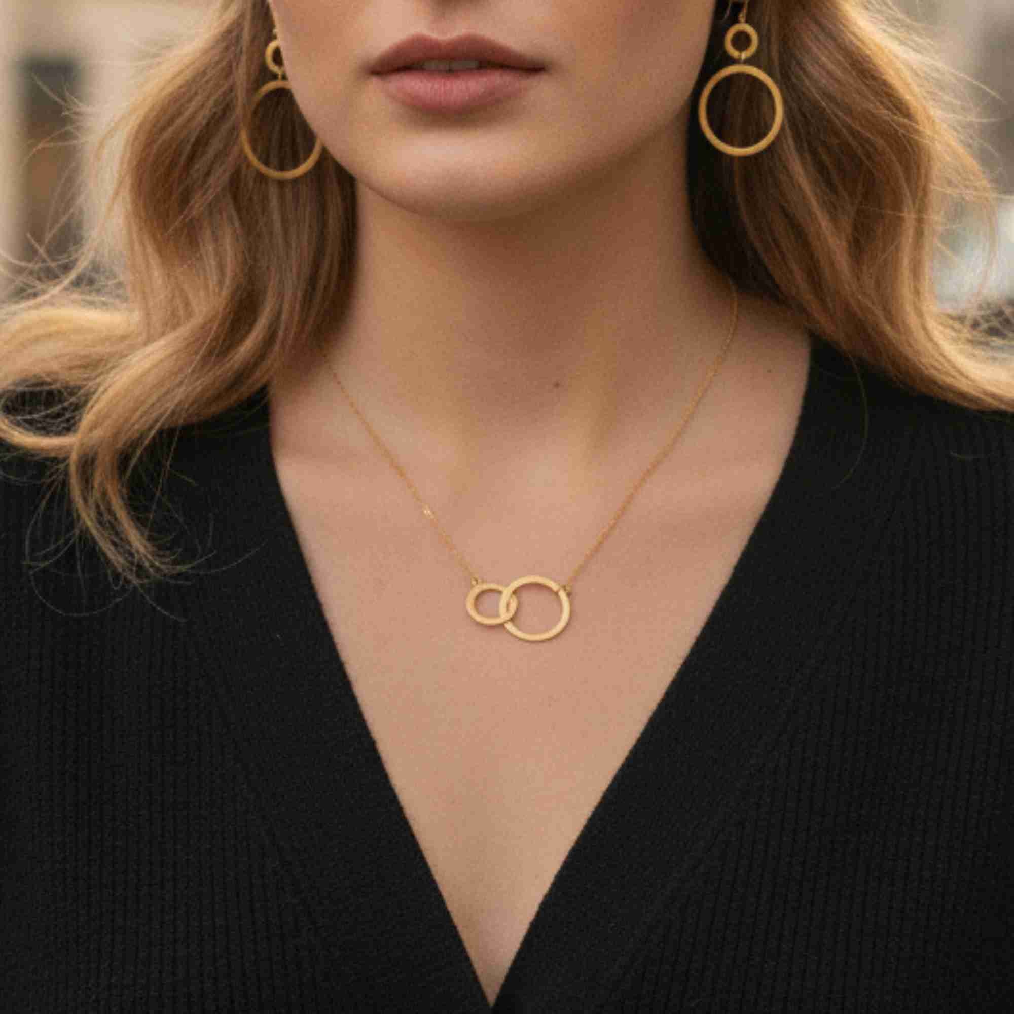 Close-up model view named Shimmering Disks (61).jpg showing a woman wearing the Shimmering Disks Interlinked Circles Necklace Gold over a black V-neck ribbed cardigan, highlighting the two delicate, interlocking gold geometric loops on a fine chain by Protsaah Zurich.