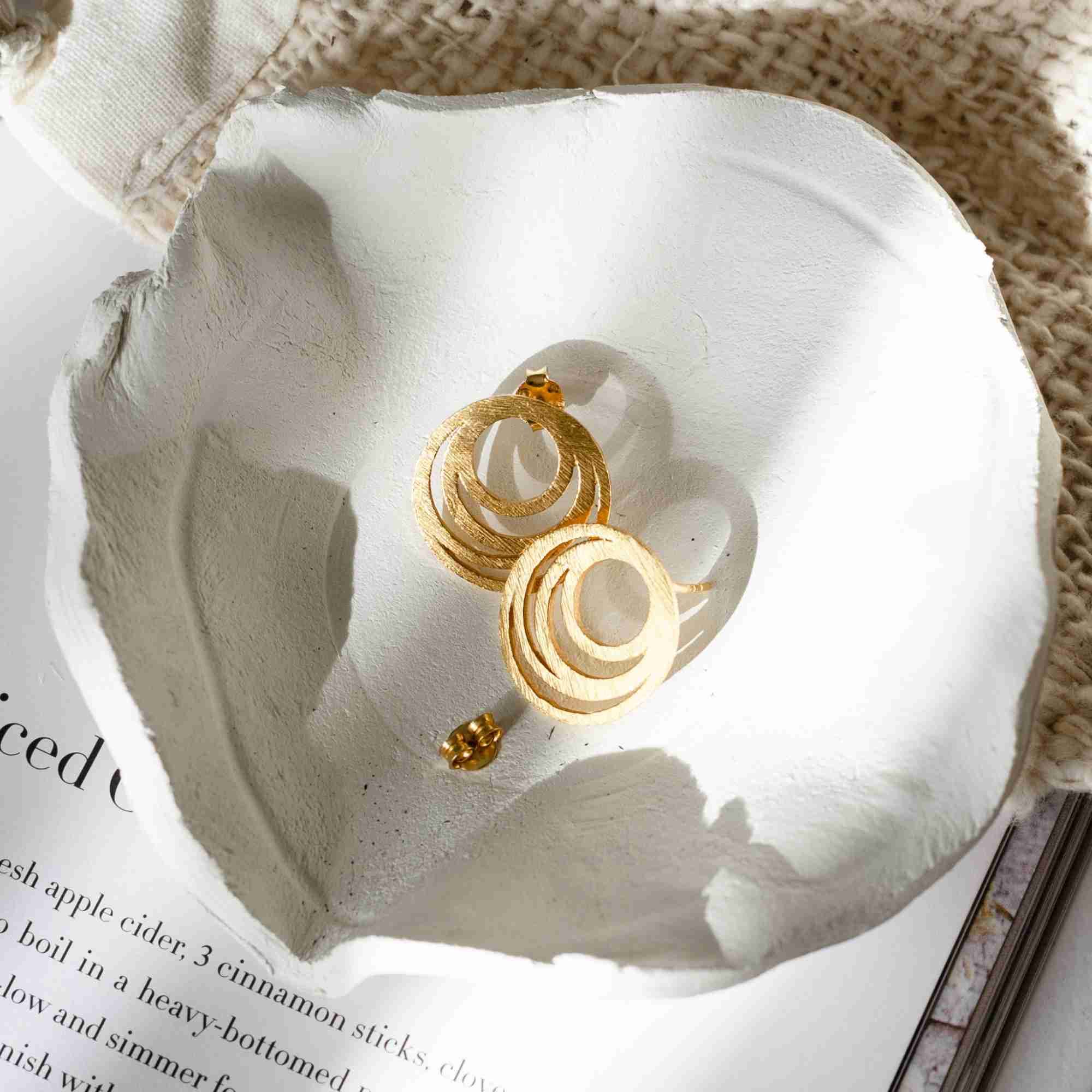 Studio product shot of Shimmering Disks Intermingled Circles Stud Earrings Gold displayed on a clean white surface, showcasing the layered concentric open-circle detailing and hand-textured matte gold finish.