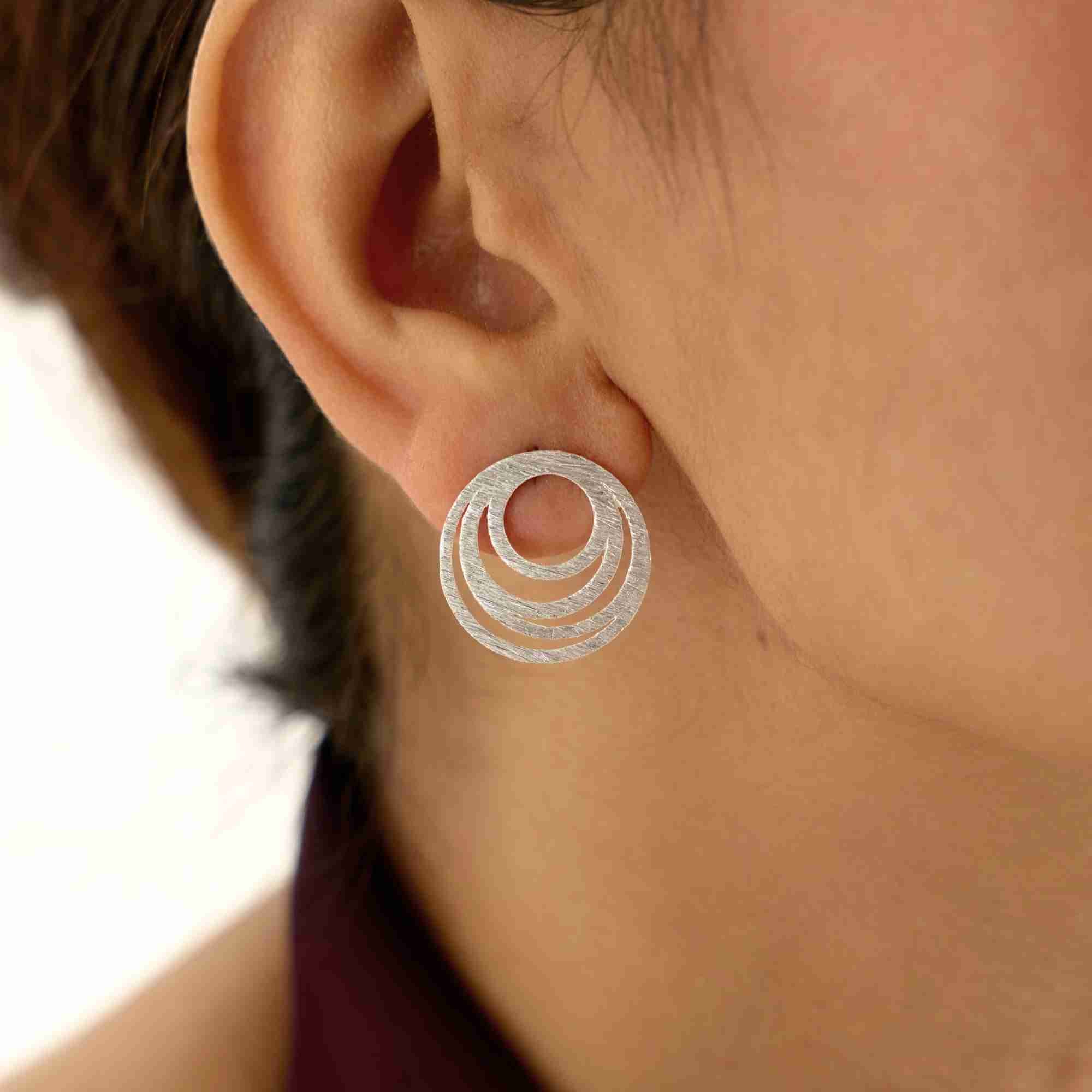 Close-up of Shimmering Disks Intermingled Circles Stud Earrings Silver, showcasing concentric circular elements and a handcrafted matte silver texture worn on the ear.