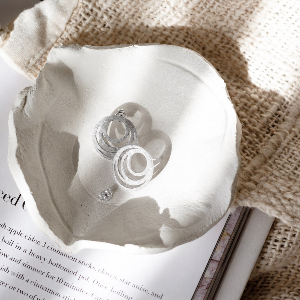 Interlocking circles stud earrings in silver with a hand-brushed finish in a white clay dish on an open book