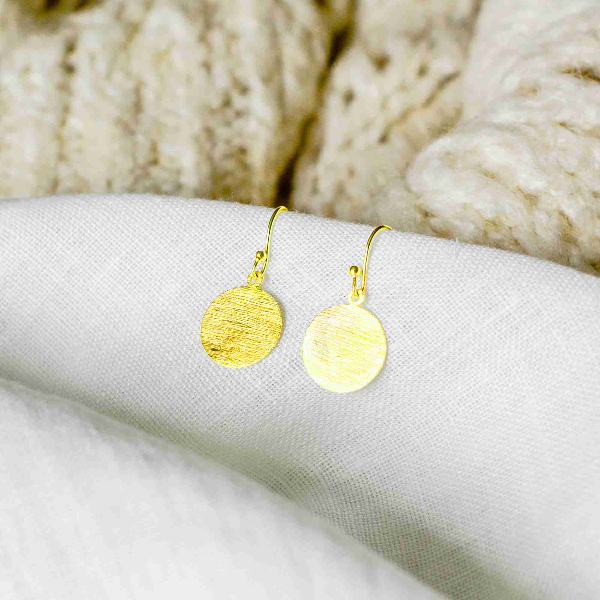 Studio product shot of Shimmering Disks Minimalist Coin Drop Earrings Medium Gold displayed flat on a textured white surface, showing the medium-sized circular coin dangles and hand-brushed matte gold texture finish.