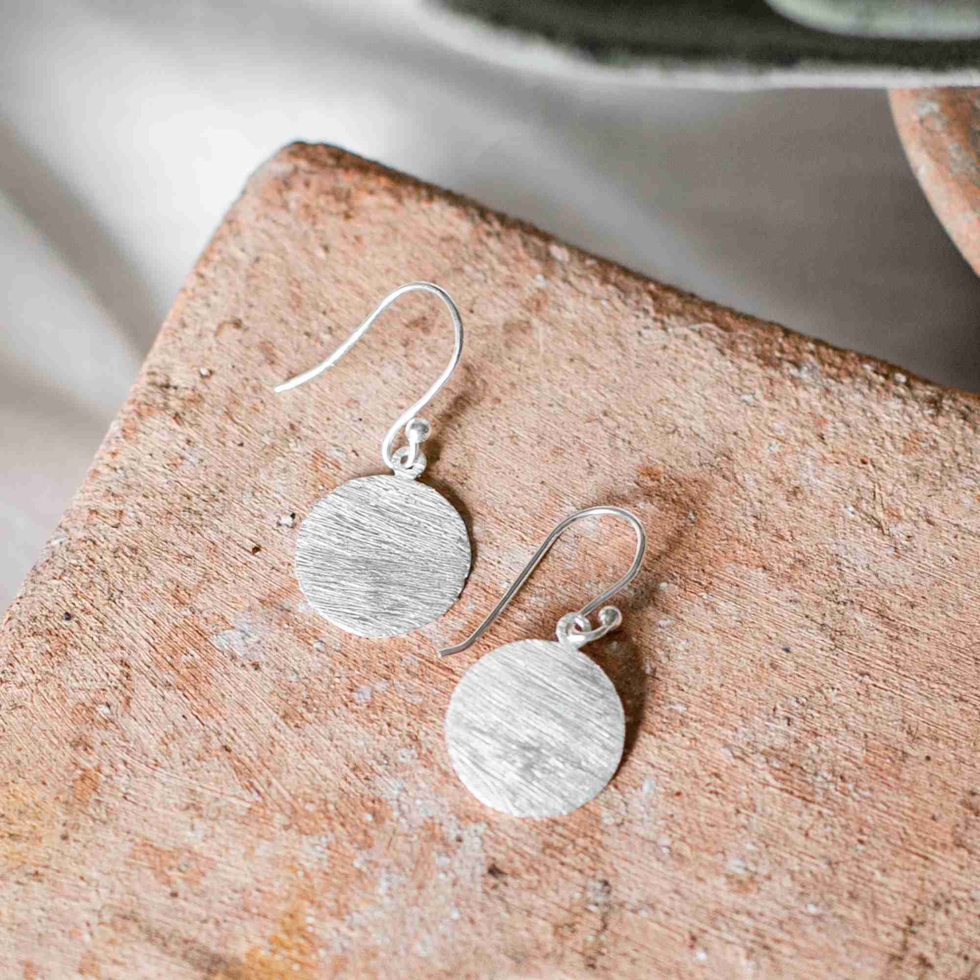 Lifestyle flatlay picture named Shimmering Disks displaying the Minimalist Coin Drop Earrings Medium Silver. A pair of handcrafted 92.5 recycled sterling silver earrings featuring flat, round disc charms with a textured, brushed metallic finish hanging from secure French hook ear wires, beautifully arranged on a rustic terracotta block by Protsaah Zurich.
