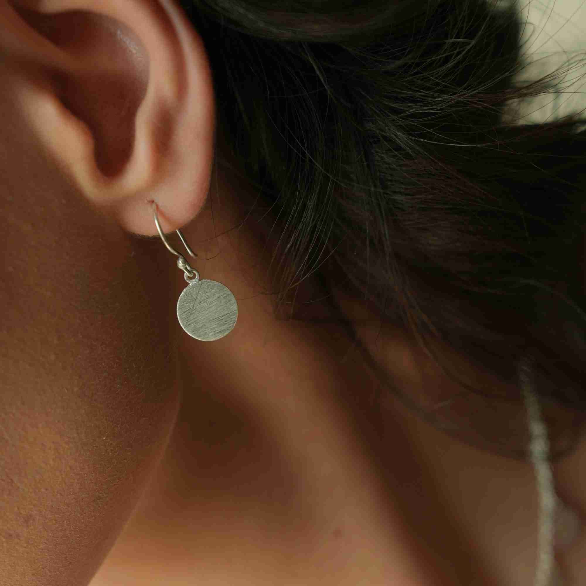 Close-up profile view of a model wearing the silver Shimmering Disks Minimalist Coin Drop Earrings Medium, showing the brushed texture on the circular disc hanging from a French ear wire hook.