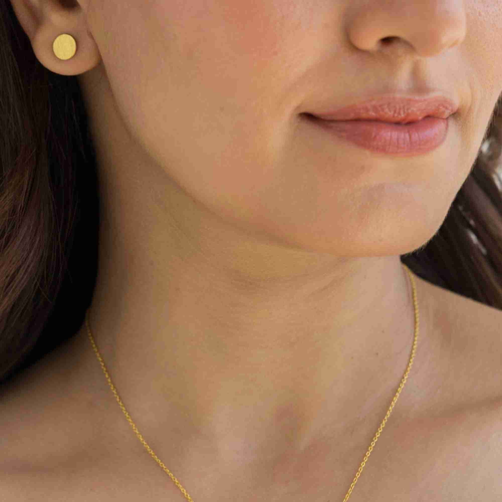 Model wearing the Shimmering Disks Minimalist Stud Earrings in 22K gold finish, showcasing the 8.2 mm flat circular disc studs on the earlobe with a fine gold chain necklace.
