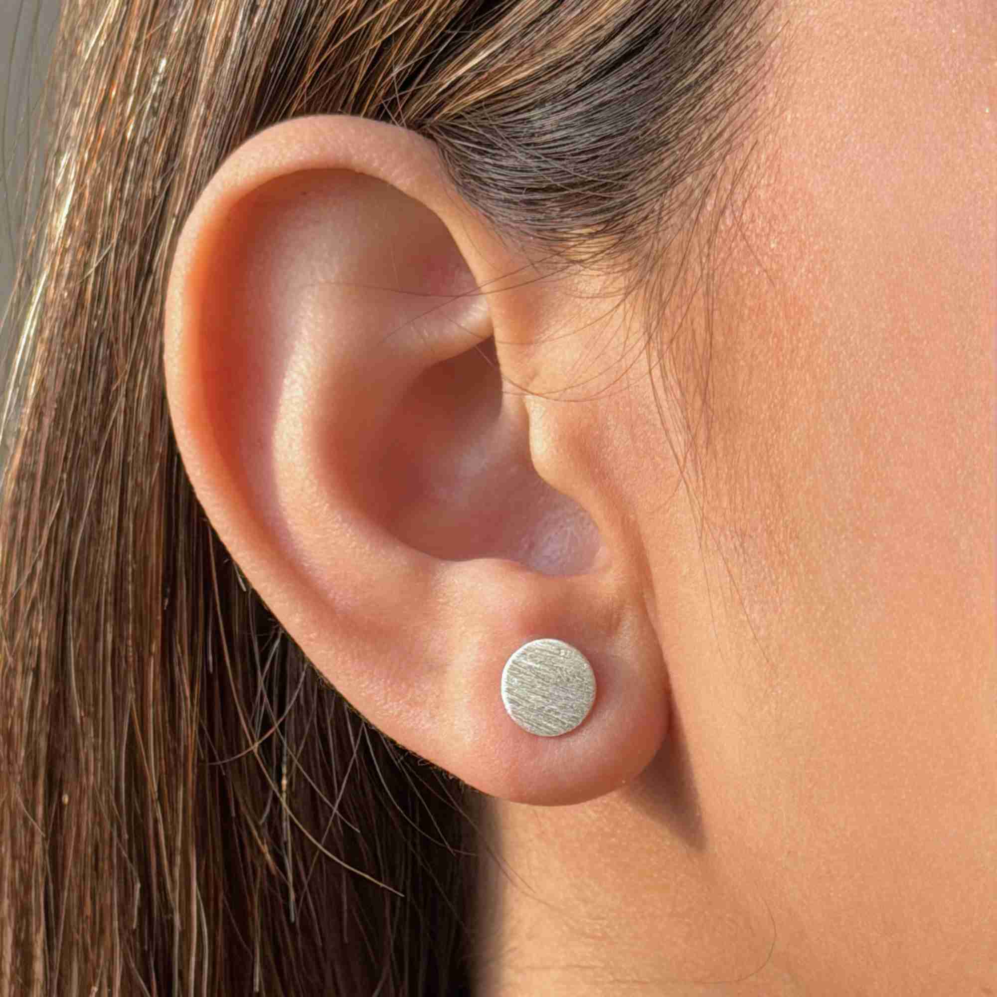 Extreme close-up of the Shimmering Disks silver circular stud earring, emphasizing the subtle matte-luster texture and round shape.