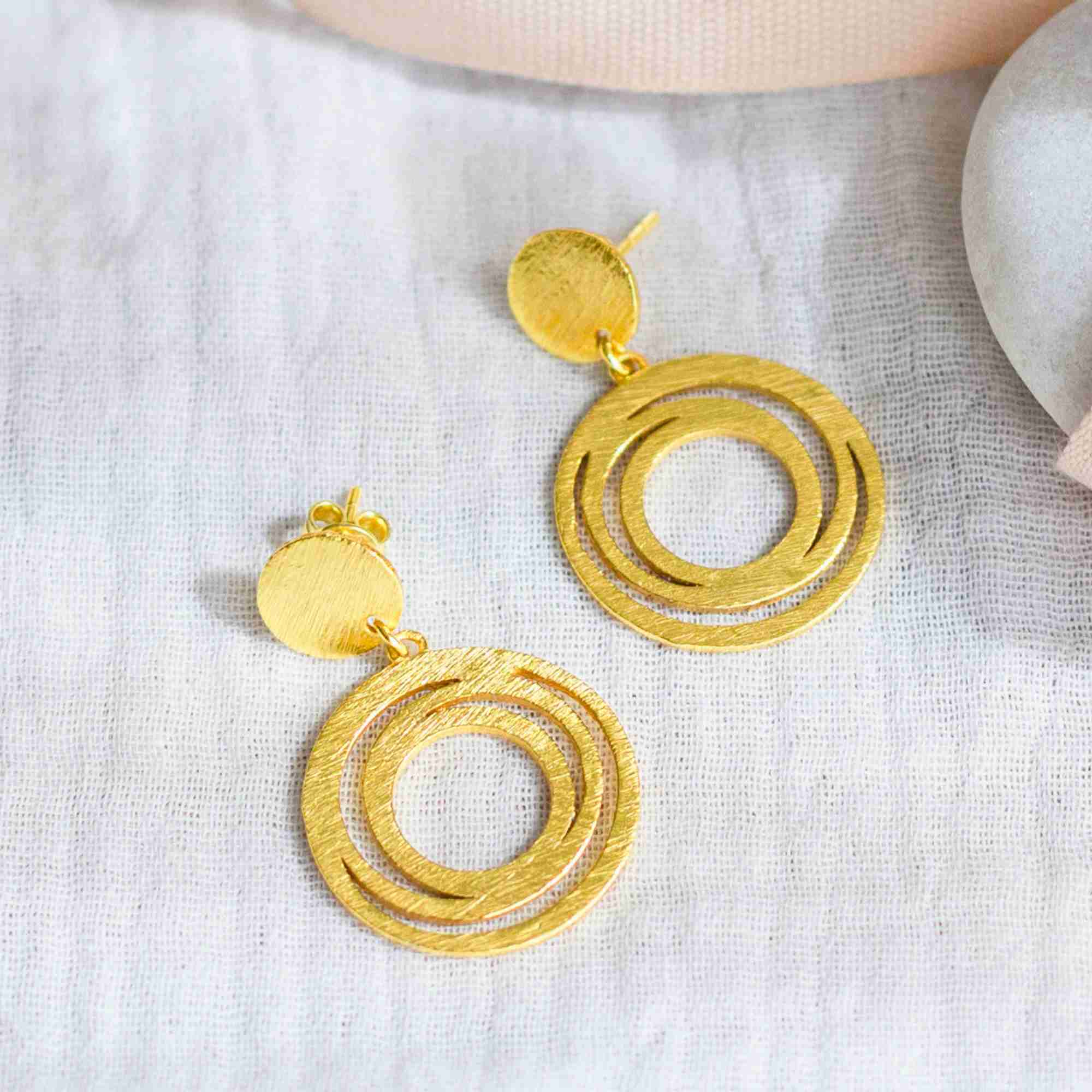 A pair of gold Shimmering Disks Orbital Swirl Drop Statement Earrings featuring round post studs and hand-brushed concentric circle swirl pendants, resting on a light linen fabric.