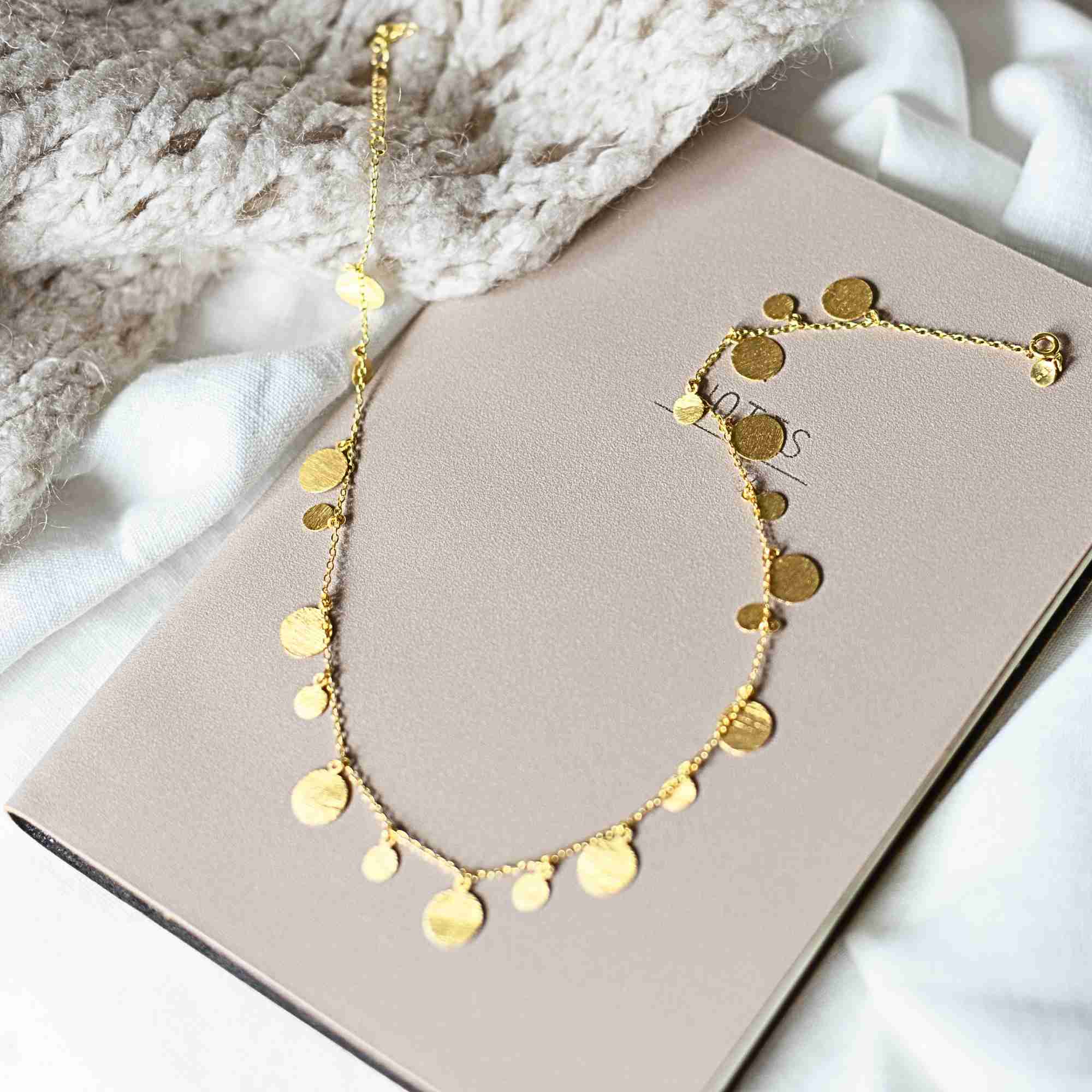 Sunlight close-up of a model wearing the gold Shimmering Disks Scattered Statement Necklace paired with a white top, black blazer, and gold statement rings.