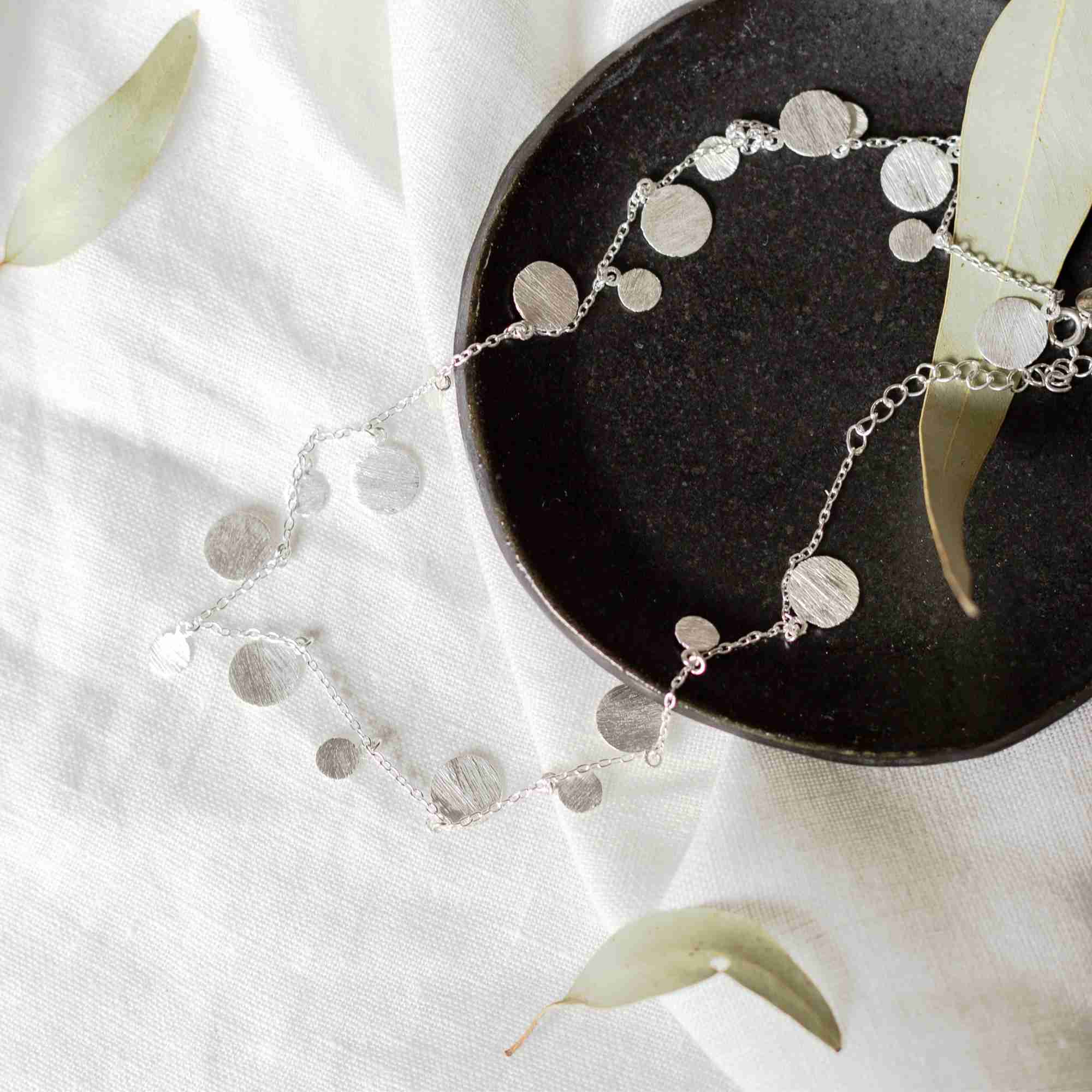 Studio flat lay view named Shimmering Disks (66).jpg showing the Shimmering Disks Scattered Statement Necklace Silver draped over a dark ceramic dish and white textured fabric with loose leaves, showcasing the multi-sized sterling silver circular charms along its fine chain by Protsaah Zurich.
