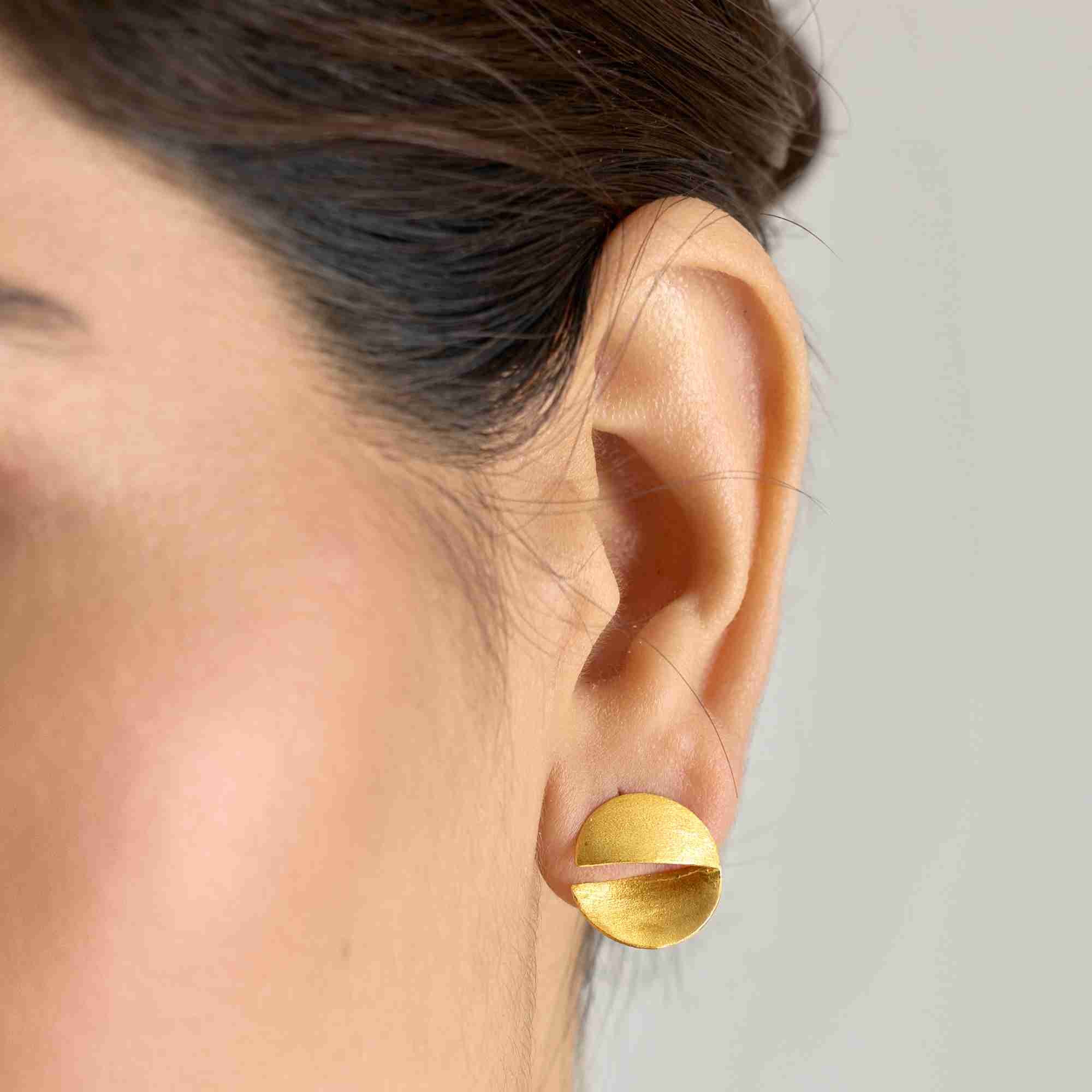 Extreme close-up of the gold Shimmering Disks Sculptural Disc Stud Earring on a model's ear, showcasing the curved, split-disc architecture and matte gold finish.