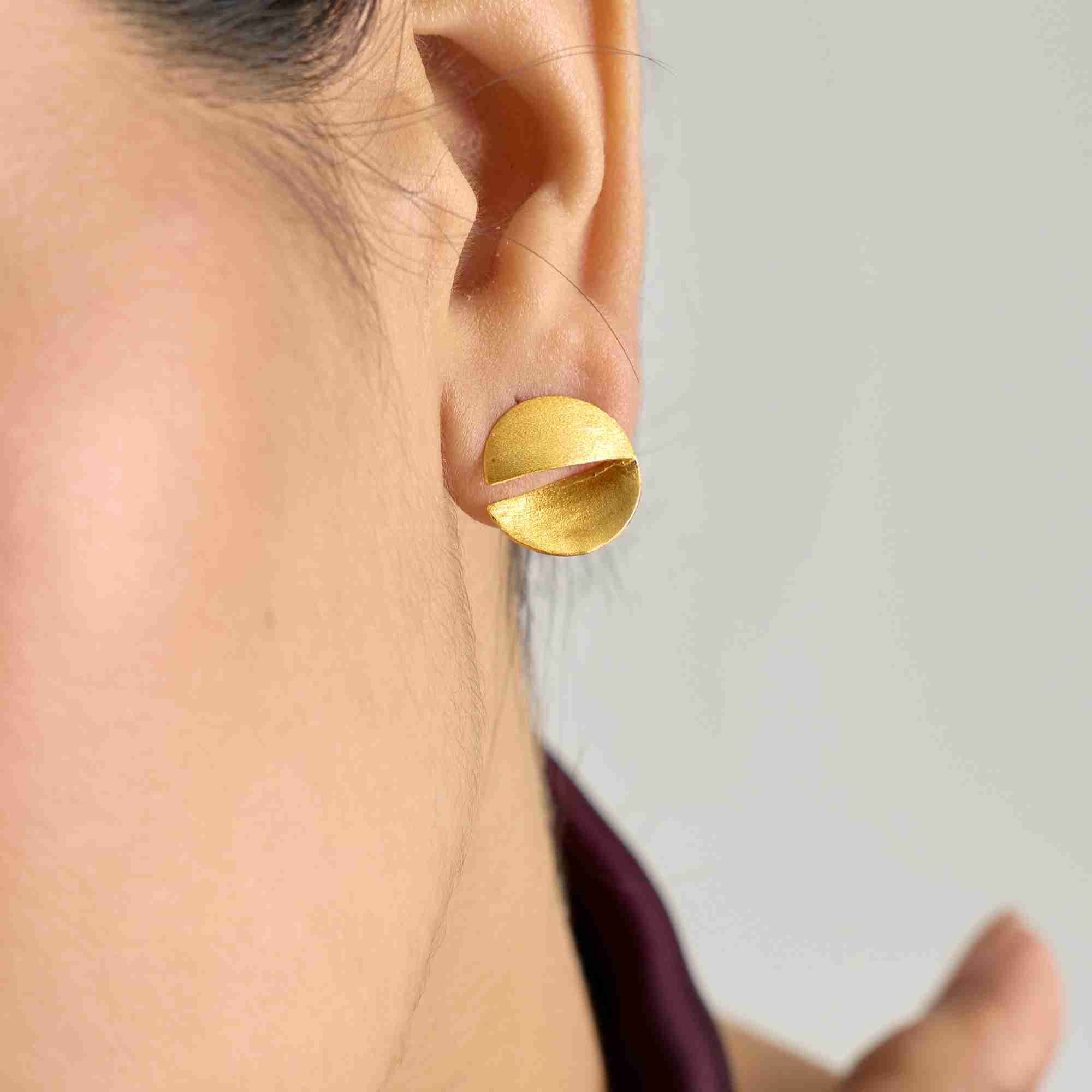 Shimmering Disks Sculptural Disc Stud Earrings Gold worn on the ear, featuring a modern circular disc design with a hand-finished matte gold texture and sculptural slit detail.